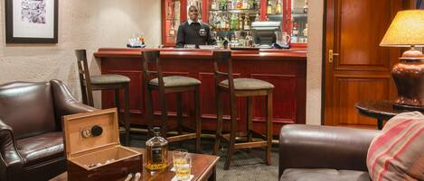 Zona de bar con un cantinero, asientos de cuero, una mesa de madera con una botella de whisky, vasos y una bandeja.