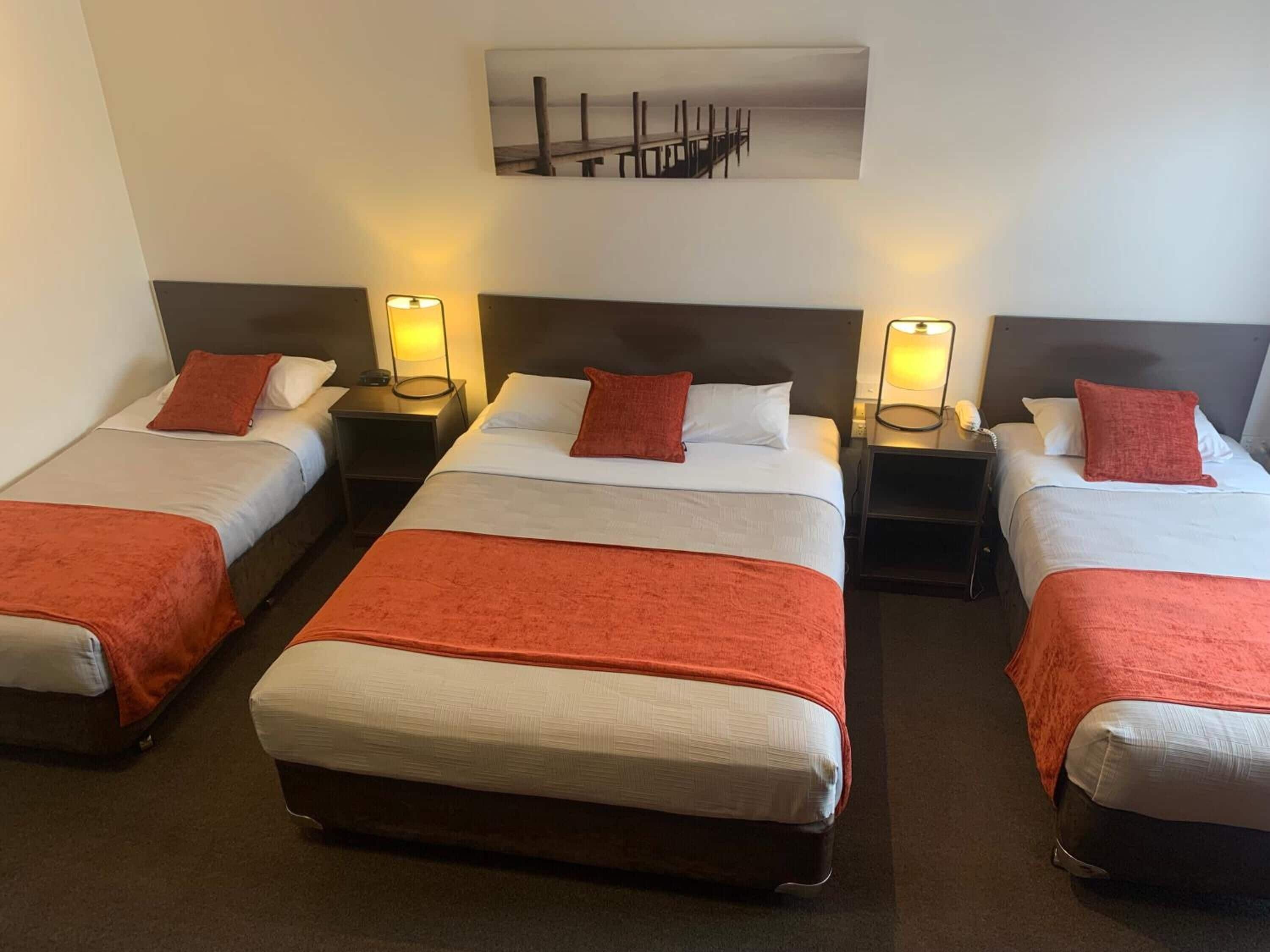 Habitación de hotel con tres camas individuales, cada una con una colcha y almohada rojas, y una cama grande con una colcha roja. Hay mesitas de noche con lámparas y un cuadro de un muelle.