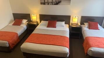 Habitación de hotel con tres camas individuales, cada una con una colcha y almohada rojas, y una cama grande con una colcha roja. Hay mesitas de noche con lámparas y un cuadro de un muelle.