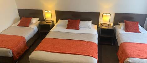 Habitación de hotel con tres camas individuales, cada una con una colcha y almohada rojas, y una cama grande con una colcha roja. Hay mesitas de noche con lámparas y un cuadro de un muelle.