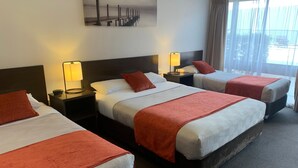 Habitación de hotel con tres camas, cada una con una colcha roja, cabecera y mesita de noche con lámpara. Se observa un cuadro en la pared sobre las camas.