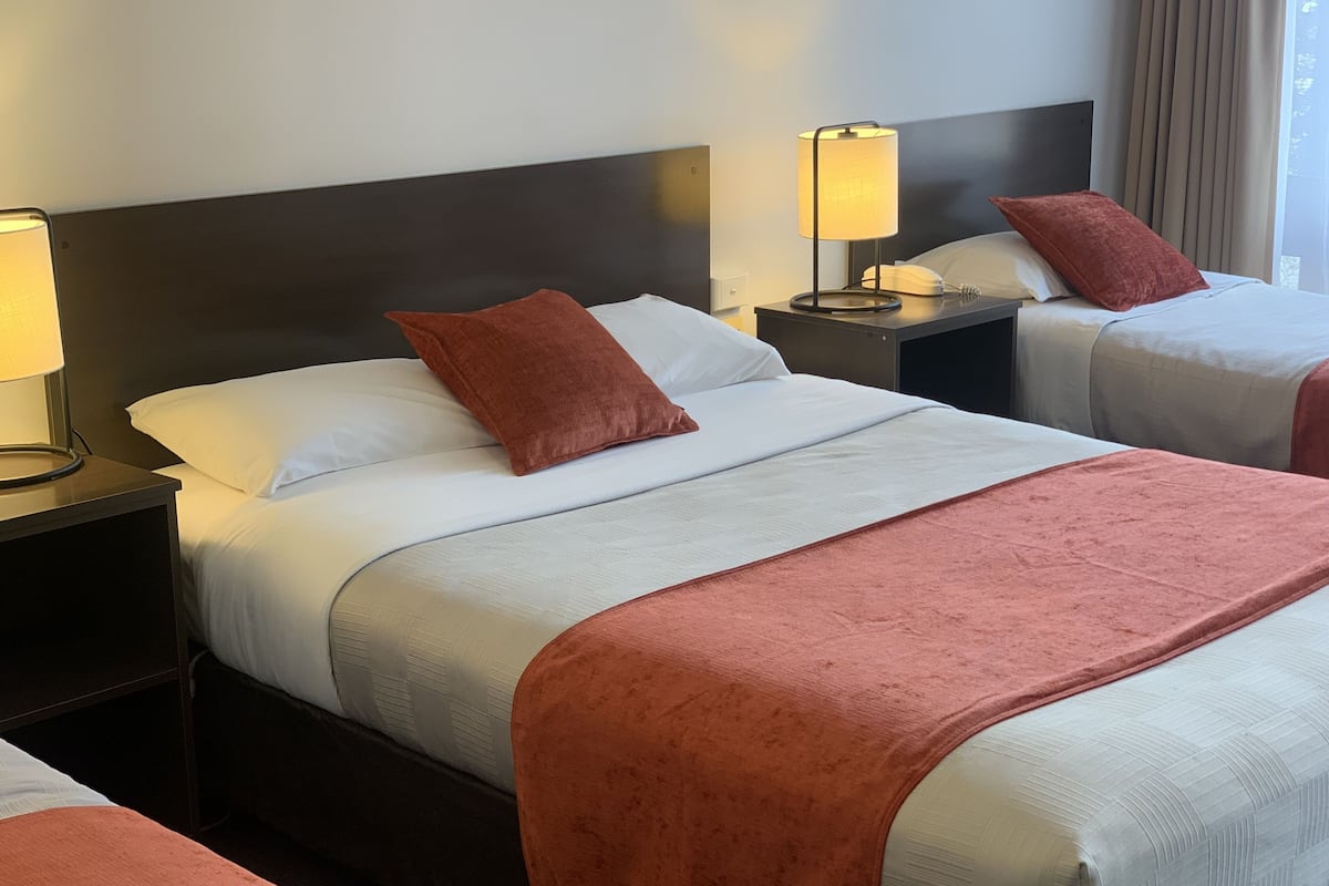 Quarto de hotel com duas camas, cada uma com roupa de cama branca e um cobertor marrom. Há duas mesas de cabeceira com abajures, e uma janela com cortinas.