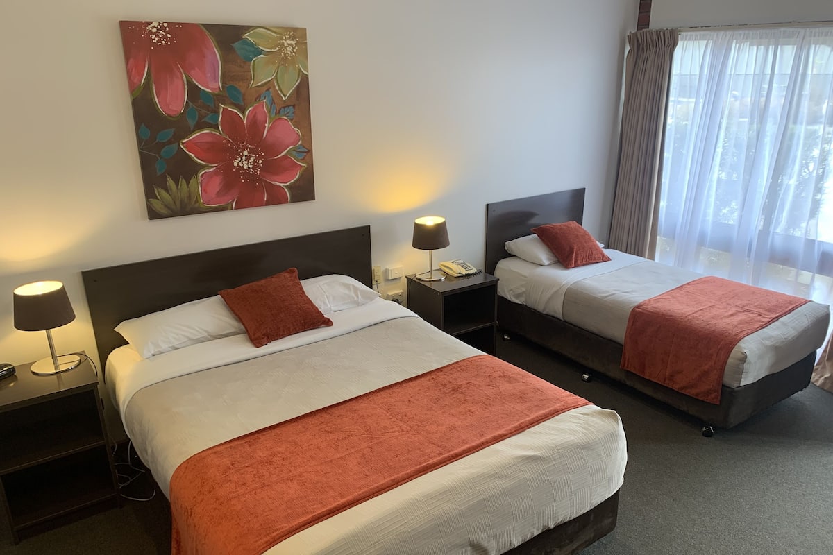 Quarto de hotel com duas camas, cada uma com um cobertor e almofada vermelhos, uma mesinha de cabeceira com luminária, um quadro na parede e uma janela com cortinas.