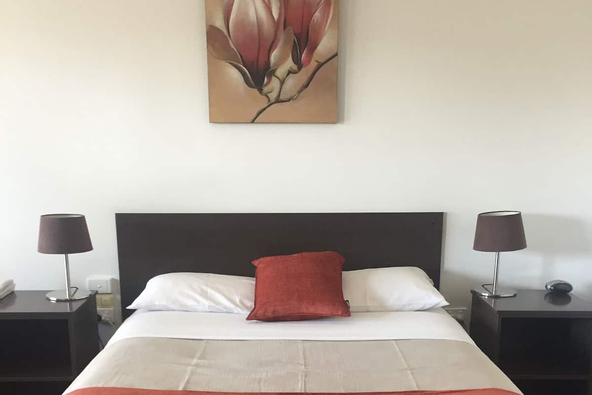 Cama bem arrumada com travesseiro vermelho, roupa de cama branca e cinza, e manta vermelha. Há duas mesas de cabeceira com luminárias de cada lado da cama.