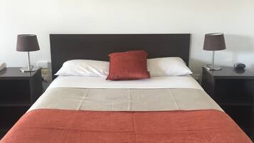 Een net opgemaakt bed met een rood kussen, wit en grijs beddengoed en een rode plaid. Aan beide zijden van het bed staan nachtkastjes met lampen.