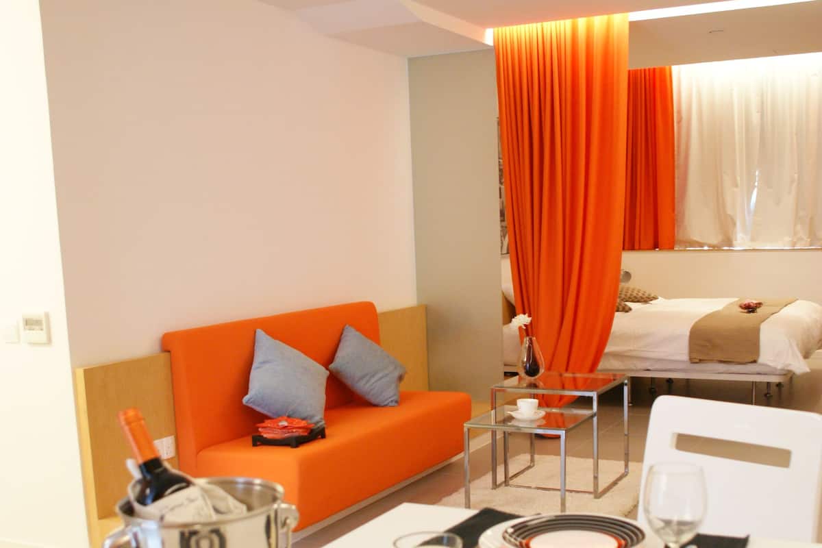 Une chambre d’hôtel avec un coin repas préparé pour un repas, un canapé et un lit agrémenté de rideaux orange.