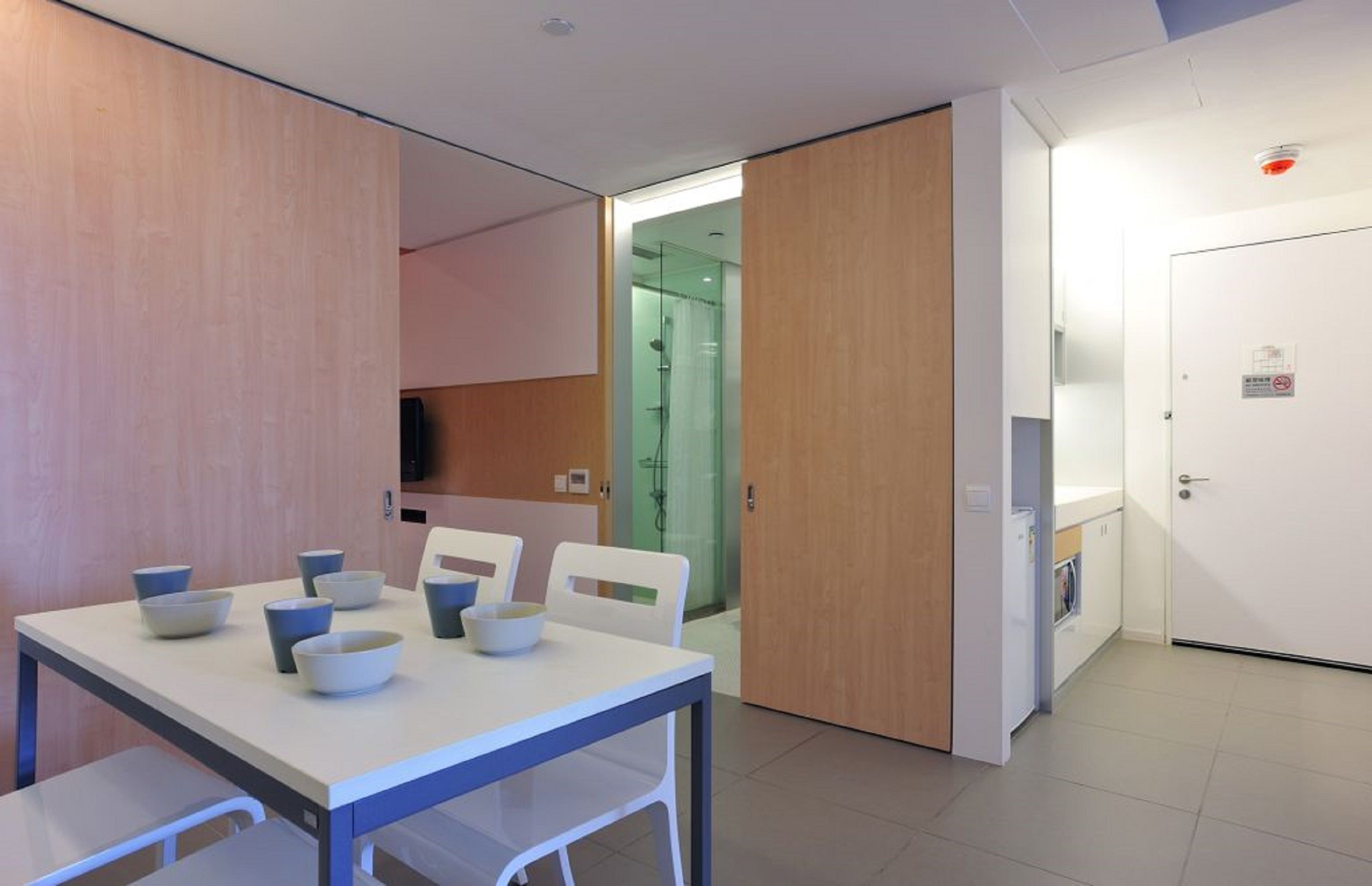 A modern hotel room with a dining area featuring a table with bowls and chairs, a shower area, and a white door with a fire alarm.