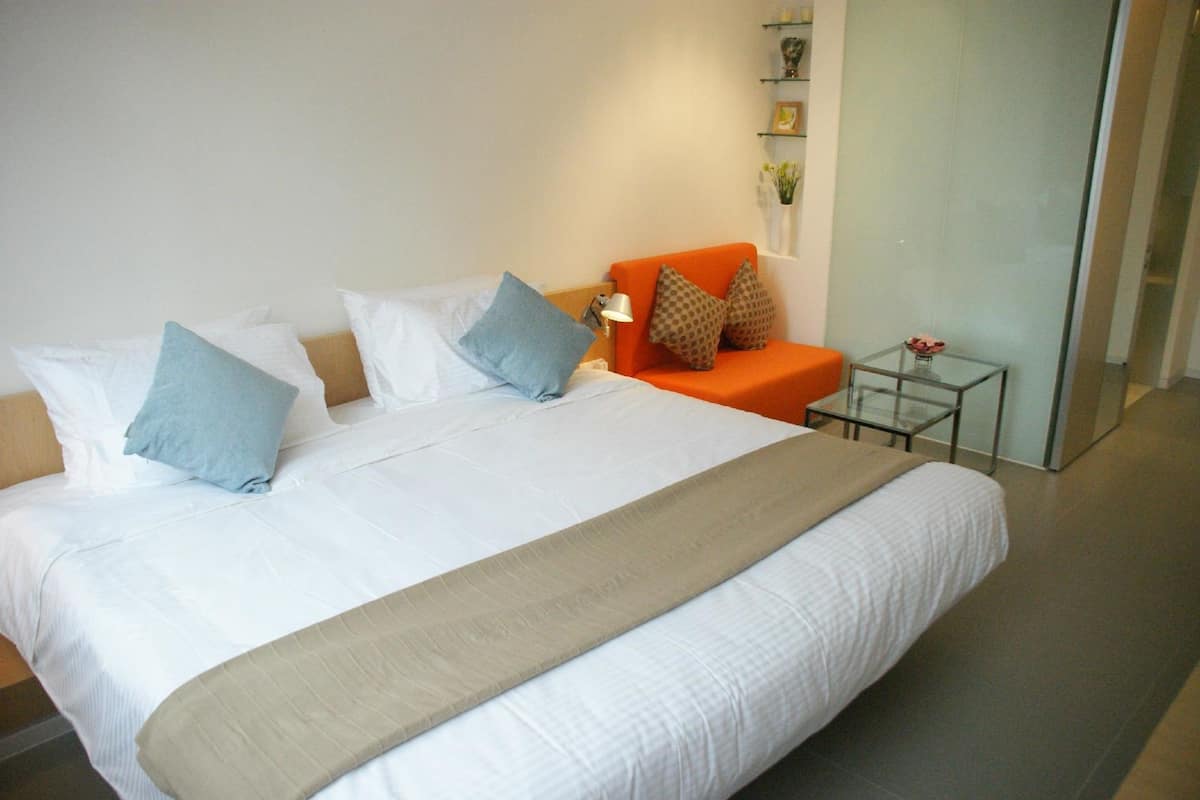 Un lit bien fait, avec du linge de lit blanc et un plaid beige, un fauteuil orange agrémenté de coussins à motifs, et une petite table en verre sur laquelle est posé un vase de fleurs.