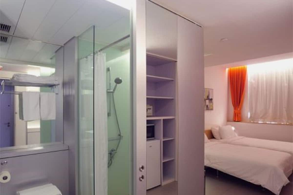 Une chambre d’hôtel moderne dotée d’une douche à parois de verre, de toilettes blanches, d’un lit avec une literie blanche et d’une armoire avec des étagères.