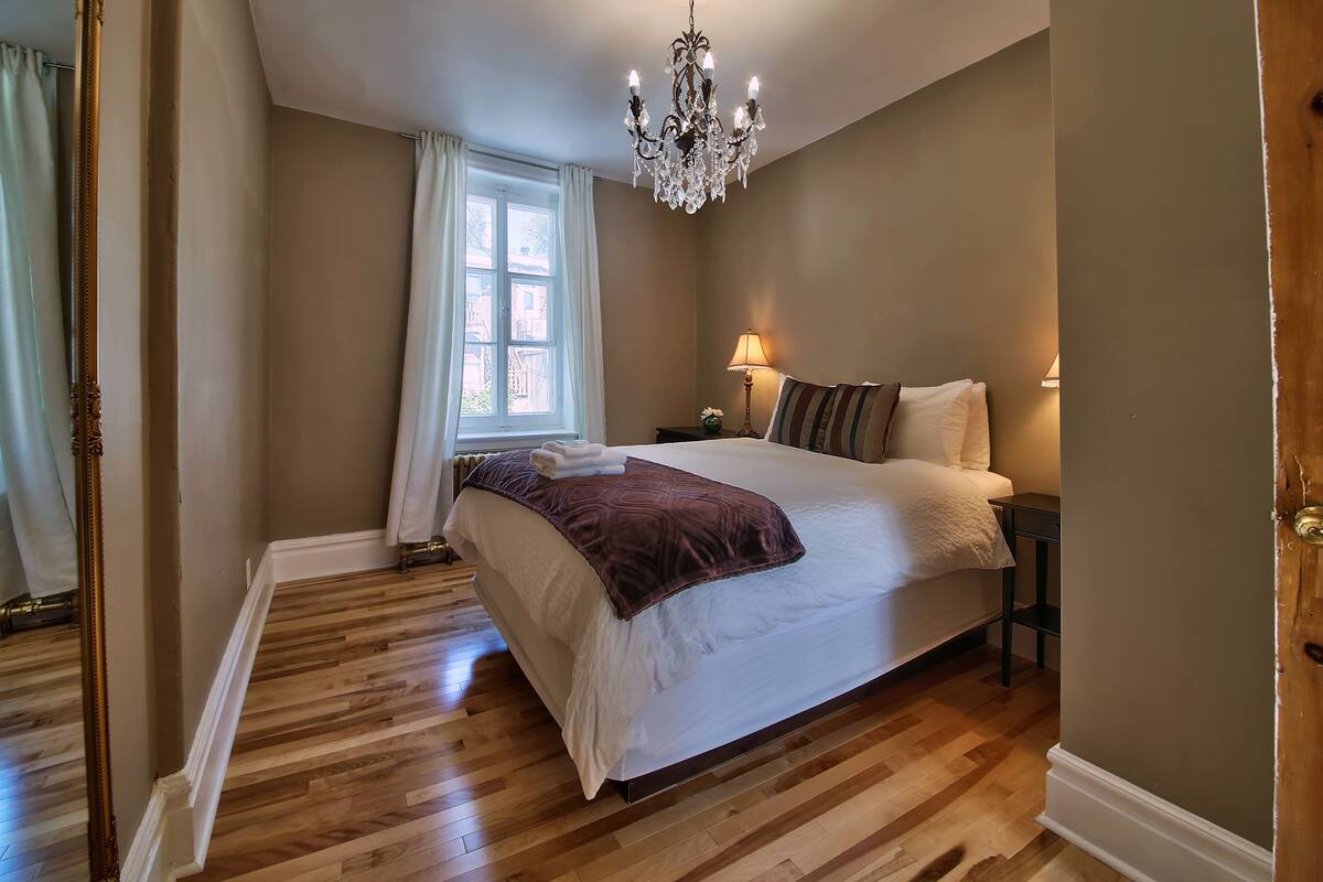 A bedroom with a large bed, a chandelier, a nightstand, and a window with curtains.