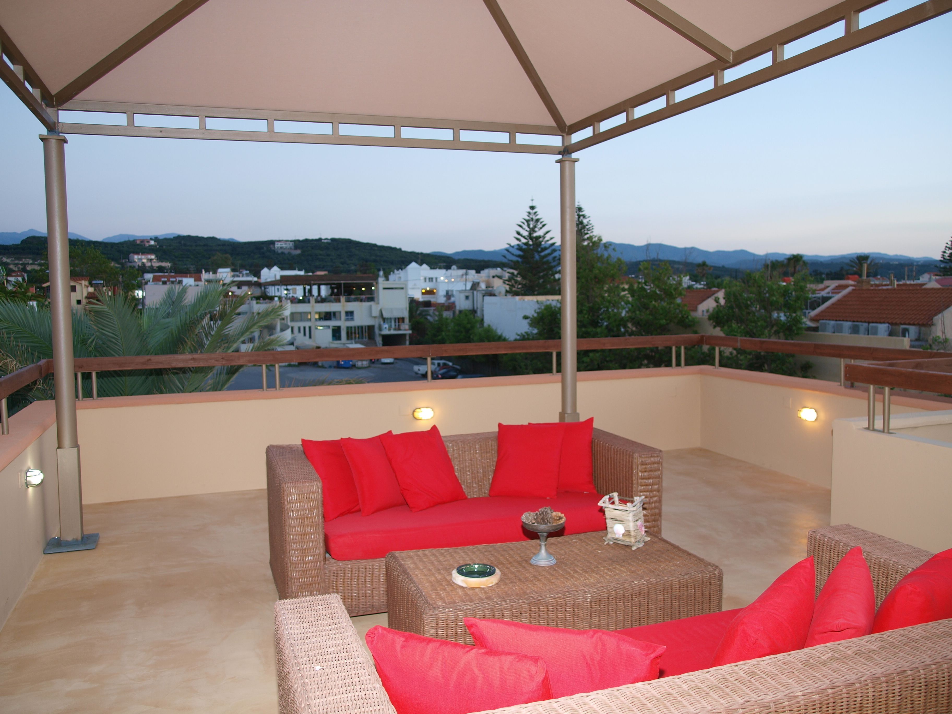 A covered outdoor seating area with wicker furniture and red cushions, overlooking a residential area with houses and mountains in the background.
