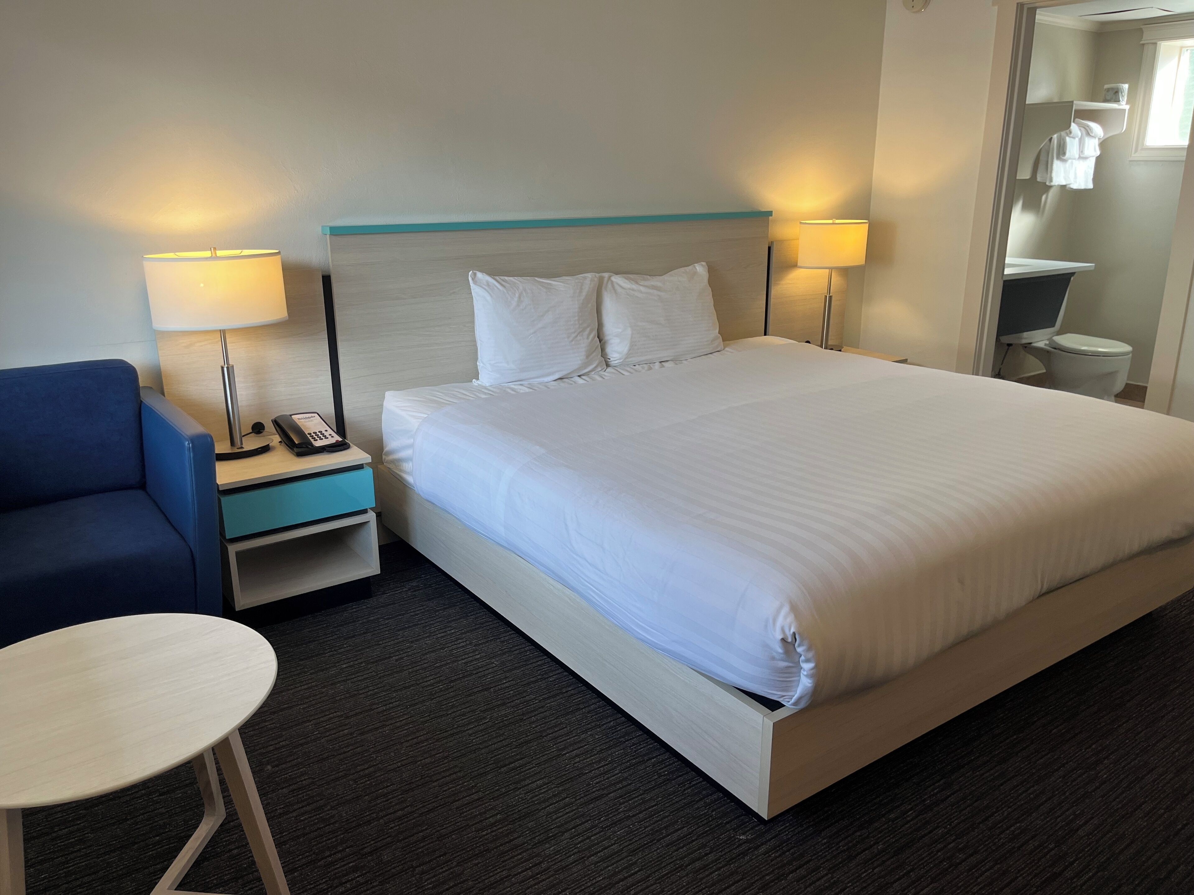 Una habitación de hotel con una cama grande, una mesita de noche con una lámpara, una silla y una pequeña mesa.