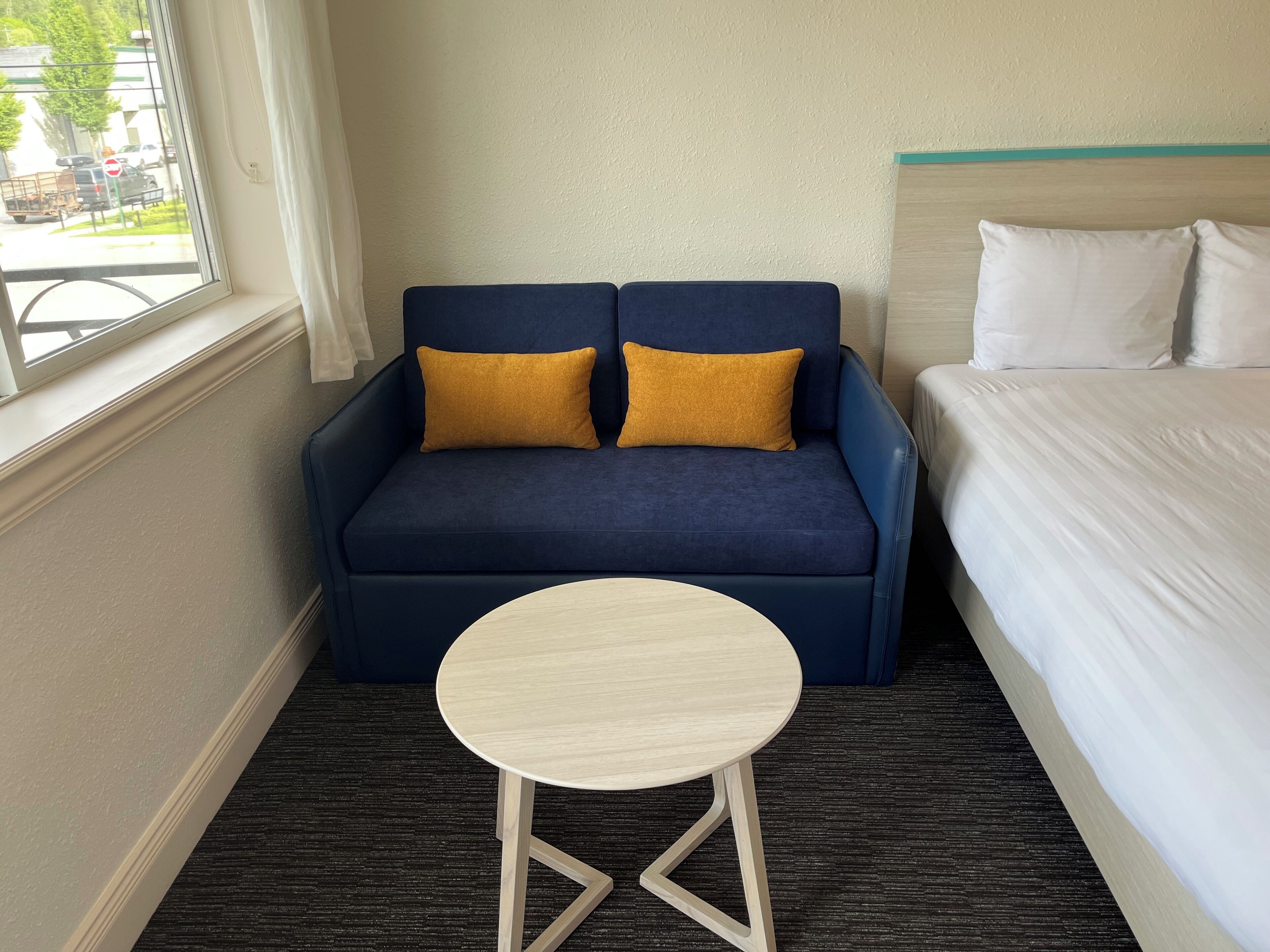 A compact hotel room with a blue sofa, two mustard pillows, a small round table, and a bed with white bedding.