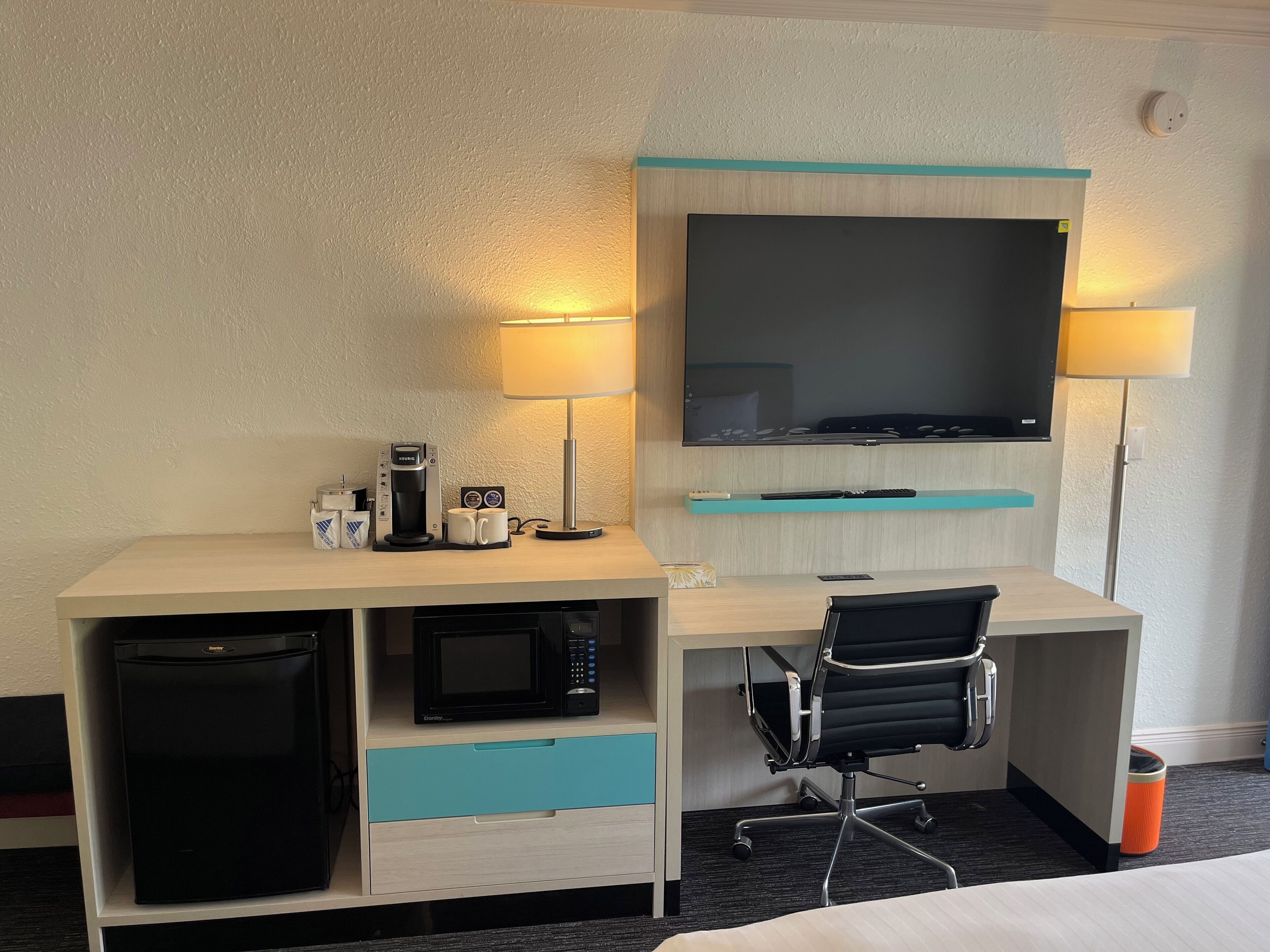 A hotel room with a desk, a television, a coffee maker, a microwave, a refrigerator, and a chair.