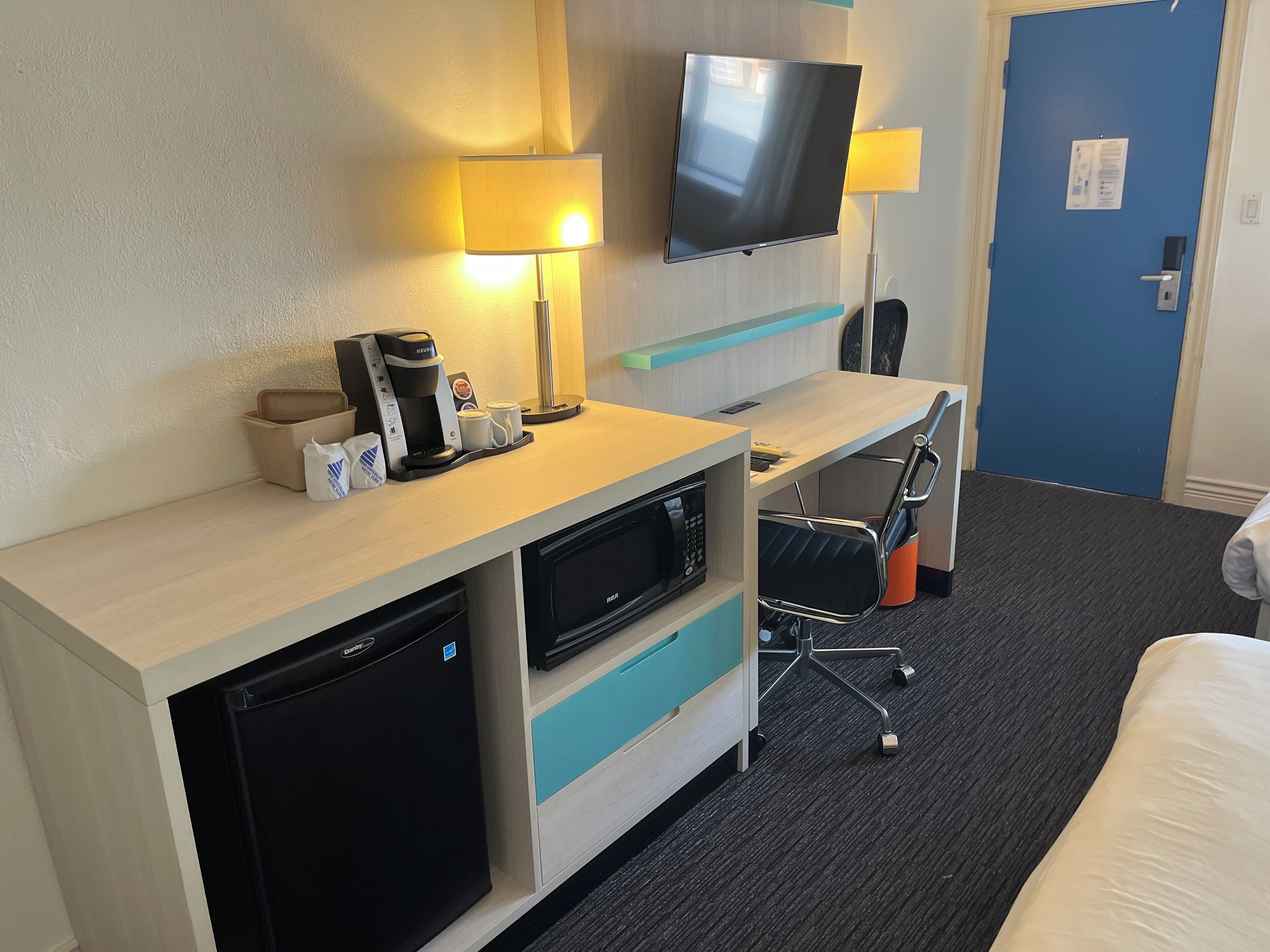 A hotel room with a bed, a desk with a coffee maker, a TV, and a microwave.