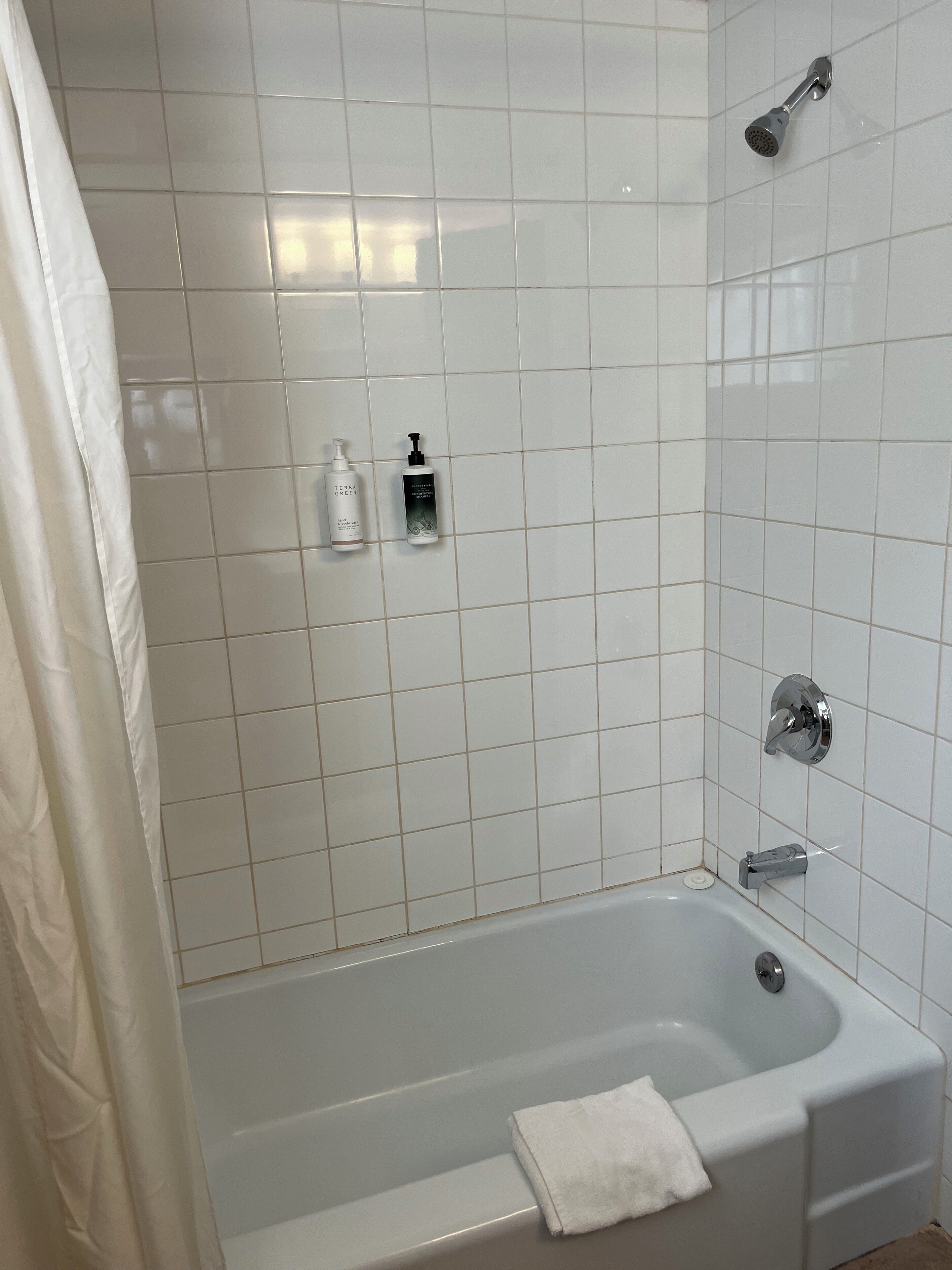 Un baño con paredes de azulejos blancos, una bañera, un cabezal de ducha y dos botellas de artículos de tocador.