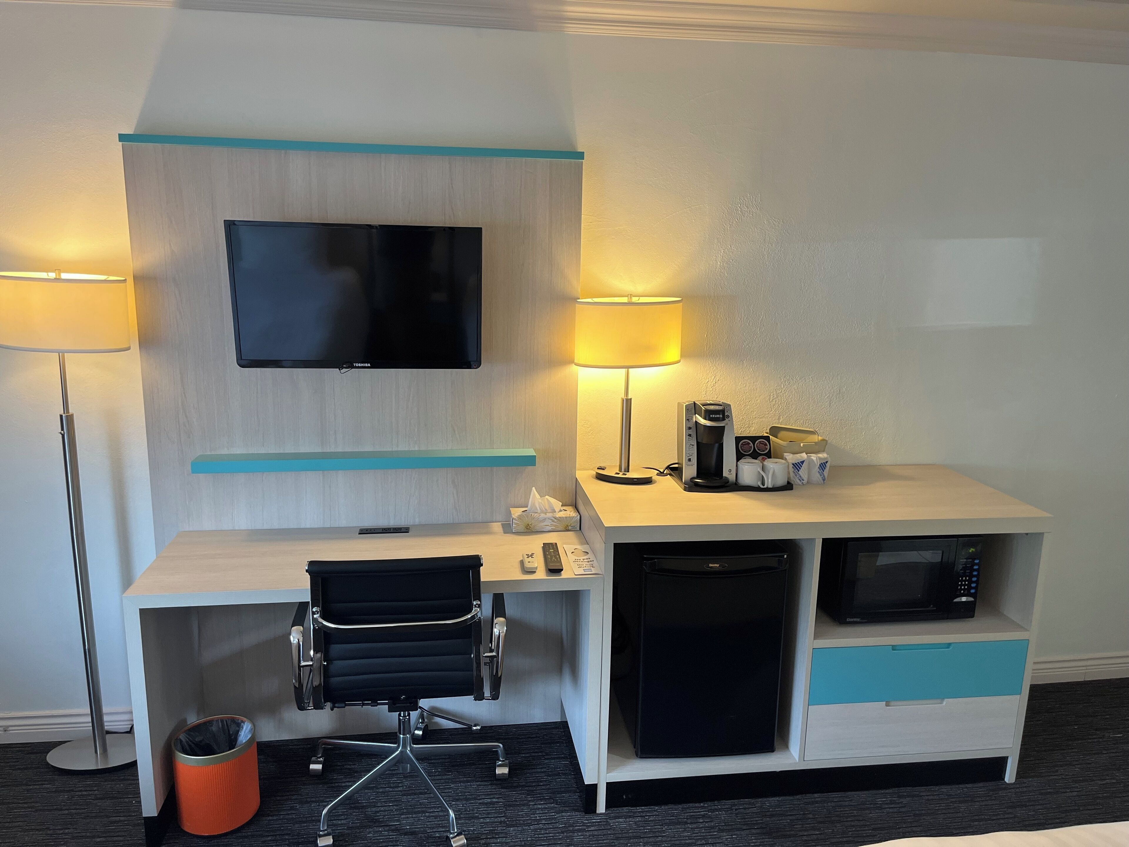 A hotel room with a desk, a flat-screen TV, a coffee maker, a microwave, and a trash can.