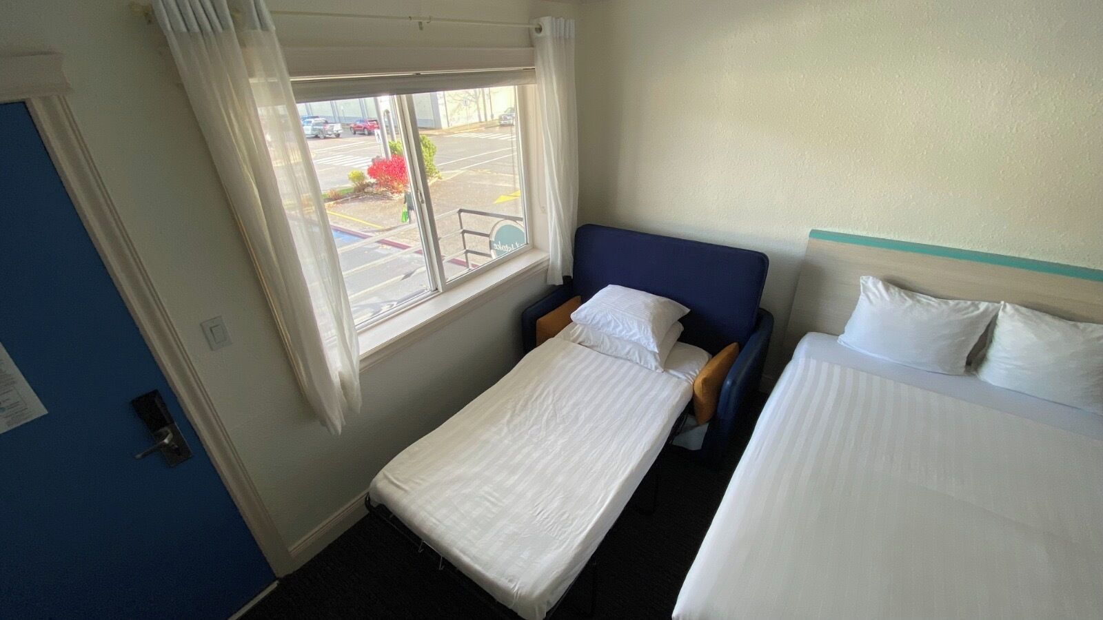 Habitación de hotel con dos camas, una ventana con cortinas, vista a un estacionamiento y una puerta con picaporte.