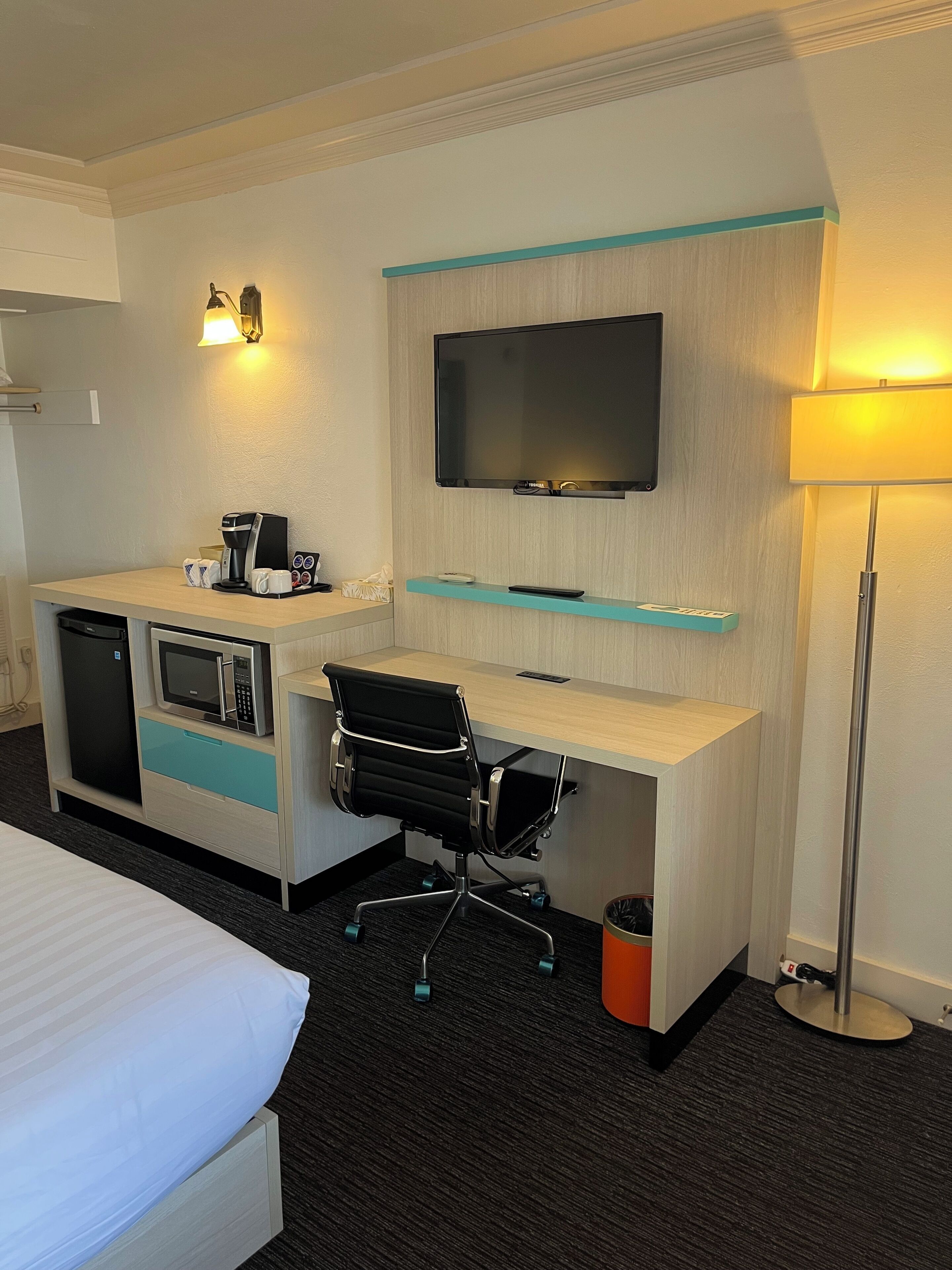 Hotellihuone, jossa on sänky, työpöytä tietokoneella, mikroaaltouuni, kahvinkeitin, televisio ja roskakori.