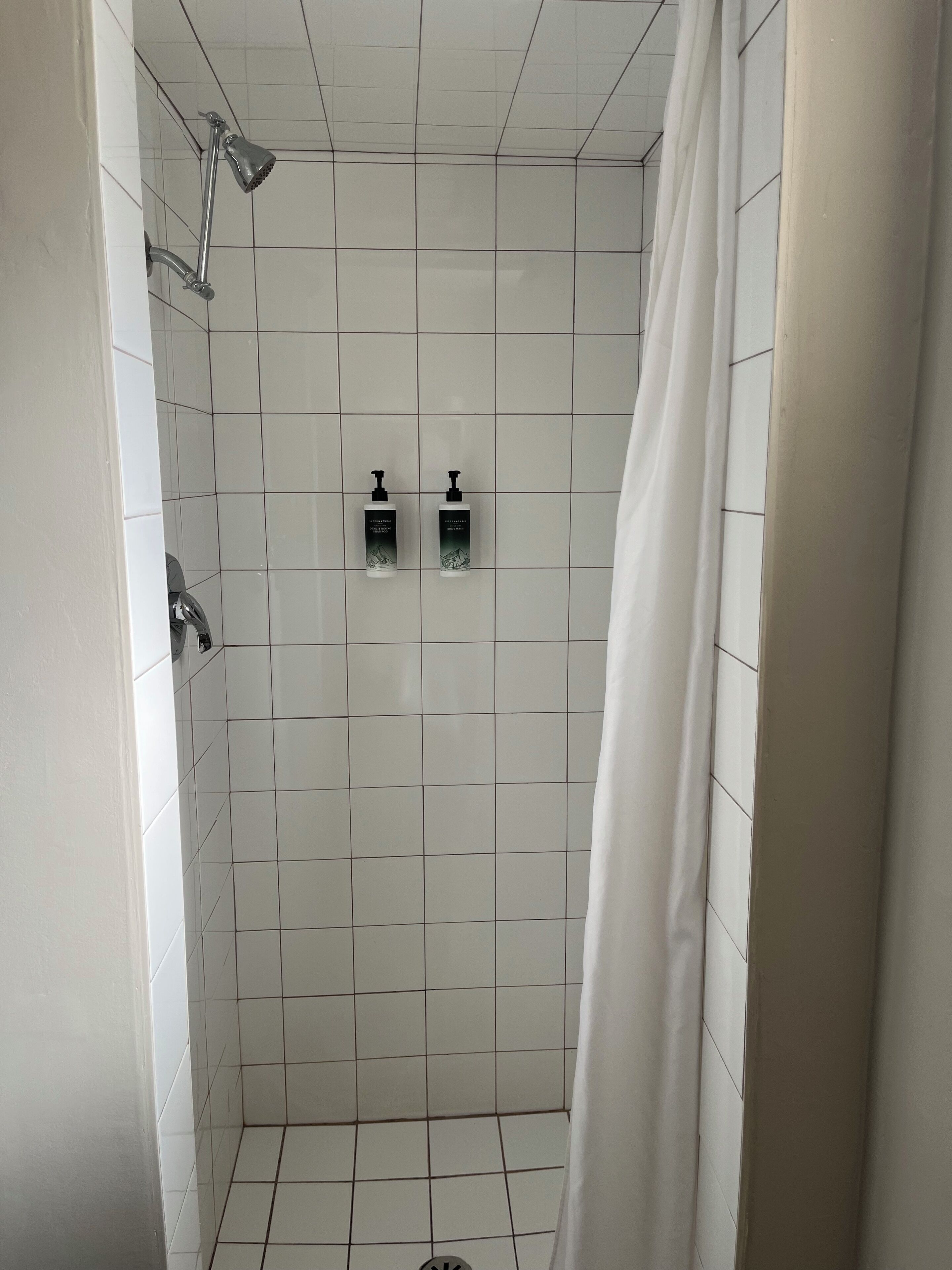 A shower area with white tiles, a showerhead, and two bottles of shampoo.