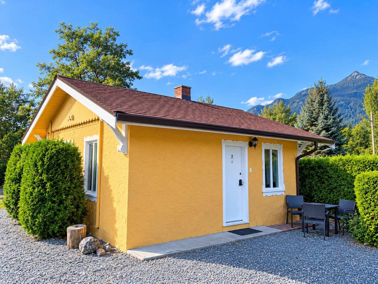 A small yellow house with a red roof, a white door, and a window. There is a gravel driveway and a small table with chairs outside.