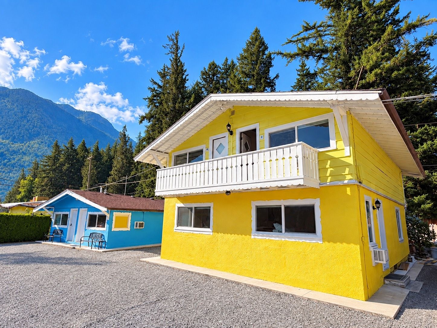 A two-story yellow building with a balcony, surrounded by trees and mountains.