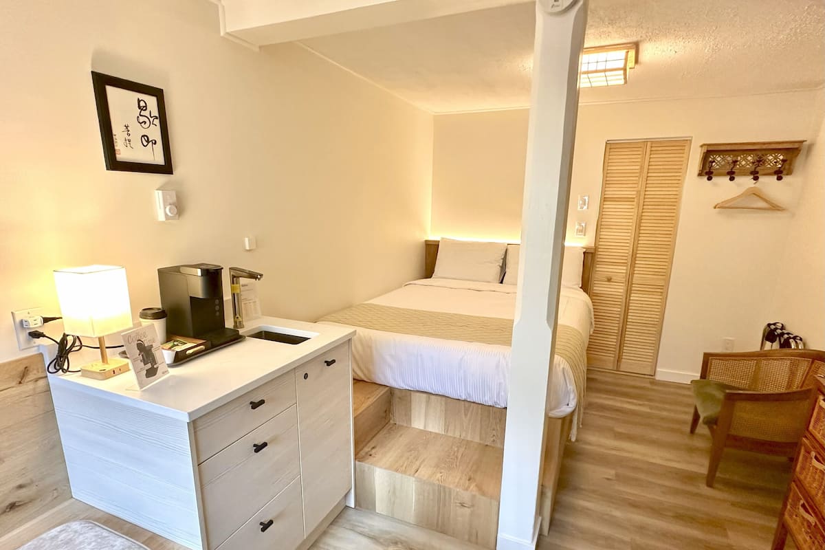 A compact hotel room with a bed, a bedside table with a lamp, a small desk with a coffee maker, a chair, and a wardrobe.