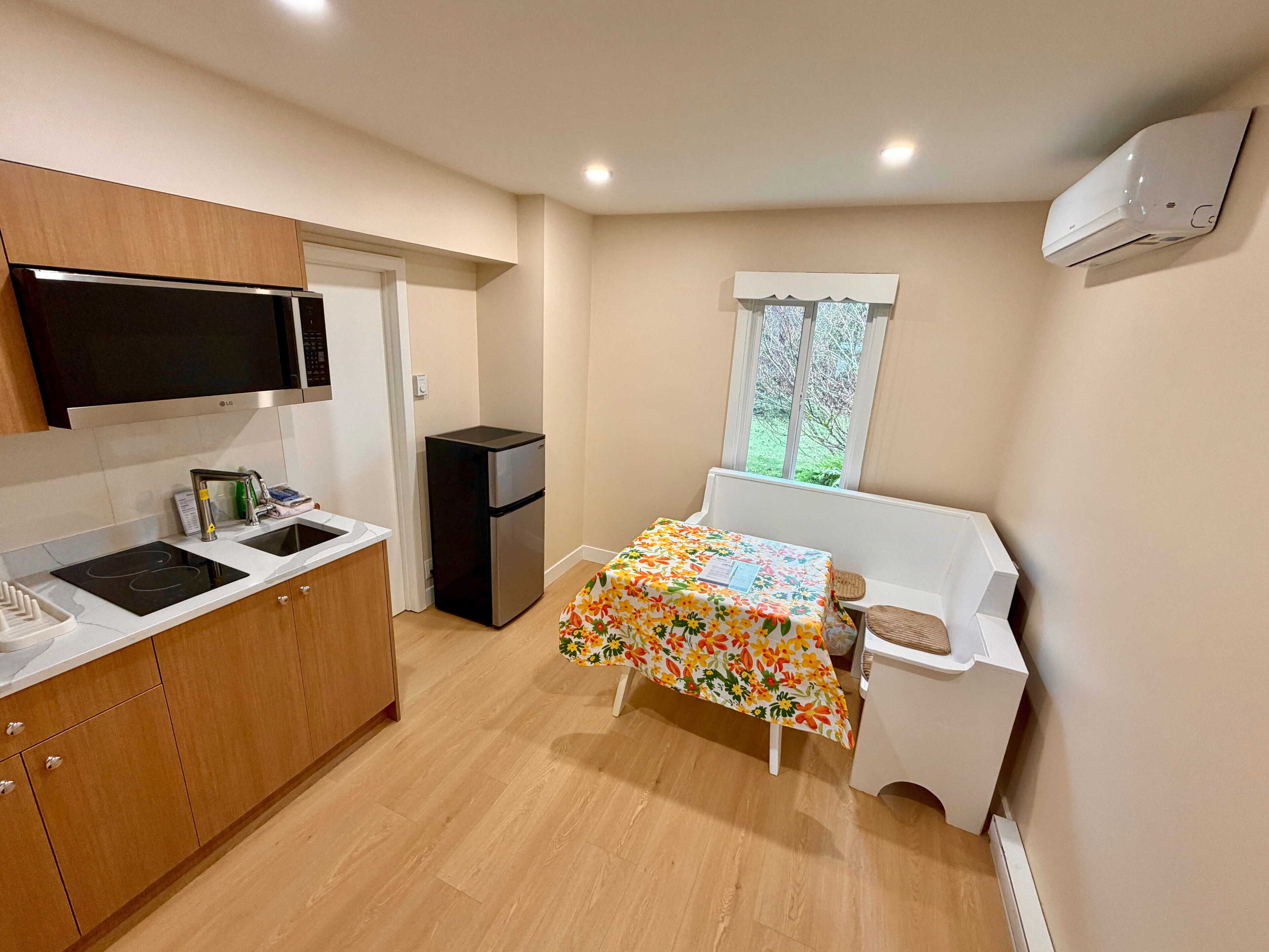 A compact kitchen with a microwave, stove, and refrigerator. There's a small table with a floral tablecloth and a chair. A window provides a view of greenery outside.