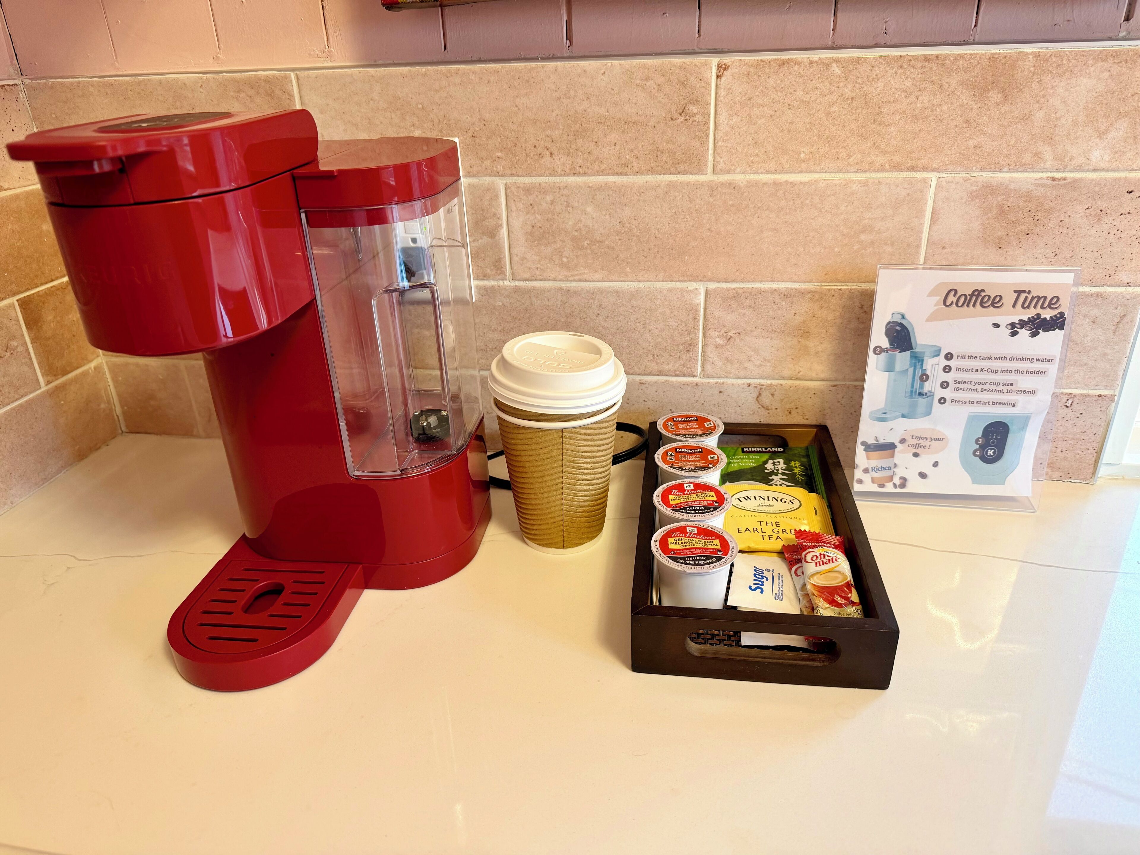 A coffee station with a red coffee maker, disposable cups, and a tray with coffee pods and a coffee machine instruction manual.