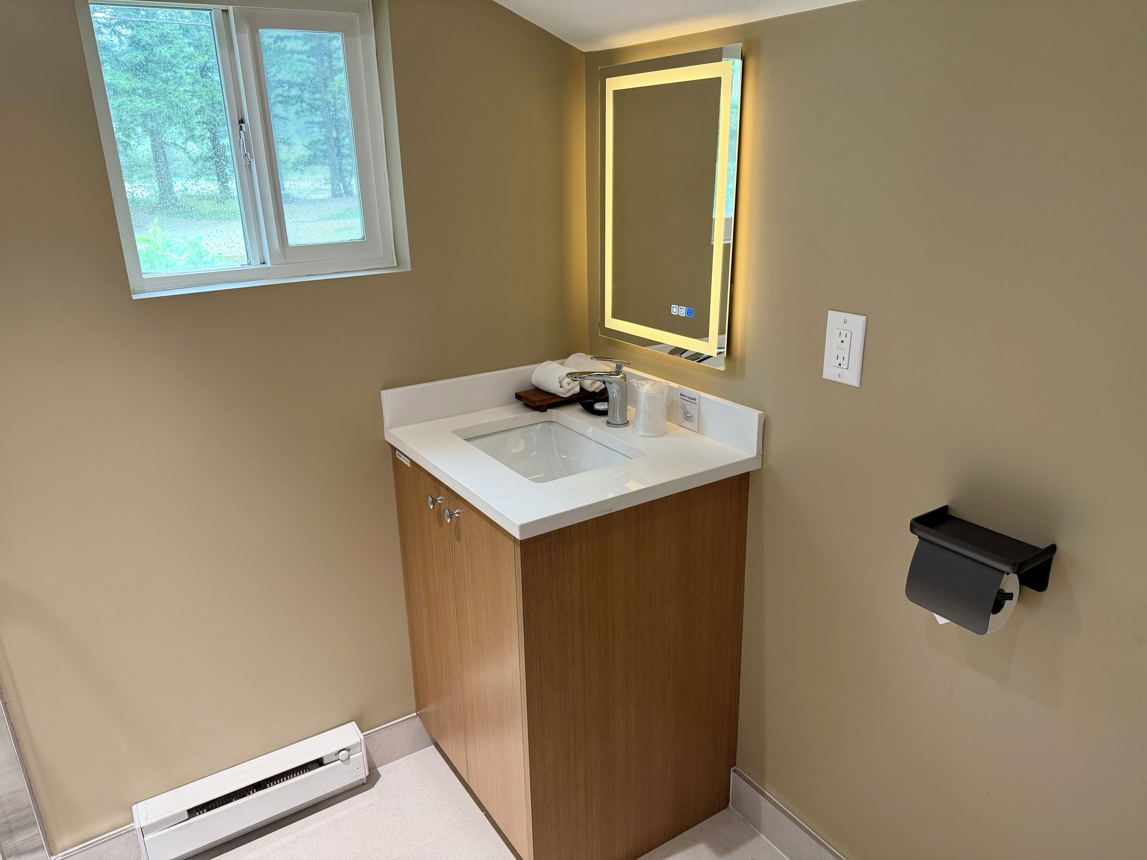 Un baño con lavamanos blanco, mueble de madera, un espejo con luces, una ventana y un perchero para toallas.