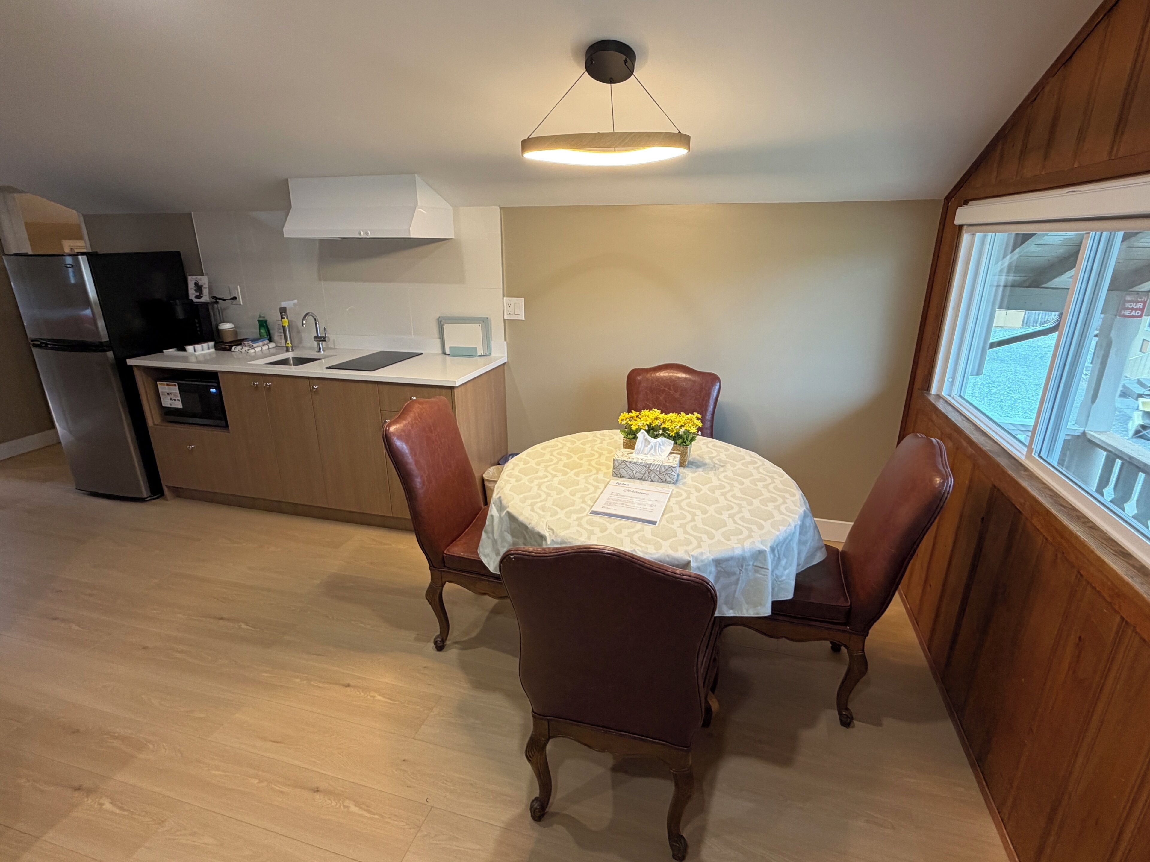Un comedor con una mesa redonda, dos sillas, un pequeño florero con flores y una cocina al fondo.