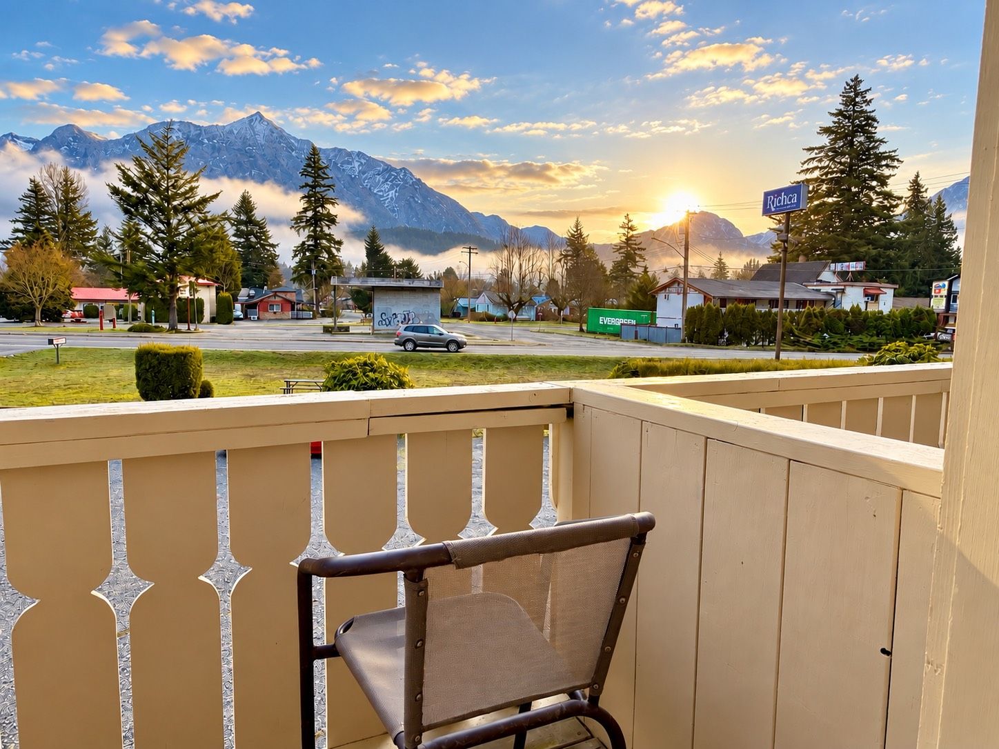 Un balcon avec une chaise, donnant sur une rue bordée de maisons, avec une voiture et des montagnes en arrière-plan.