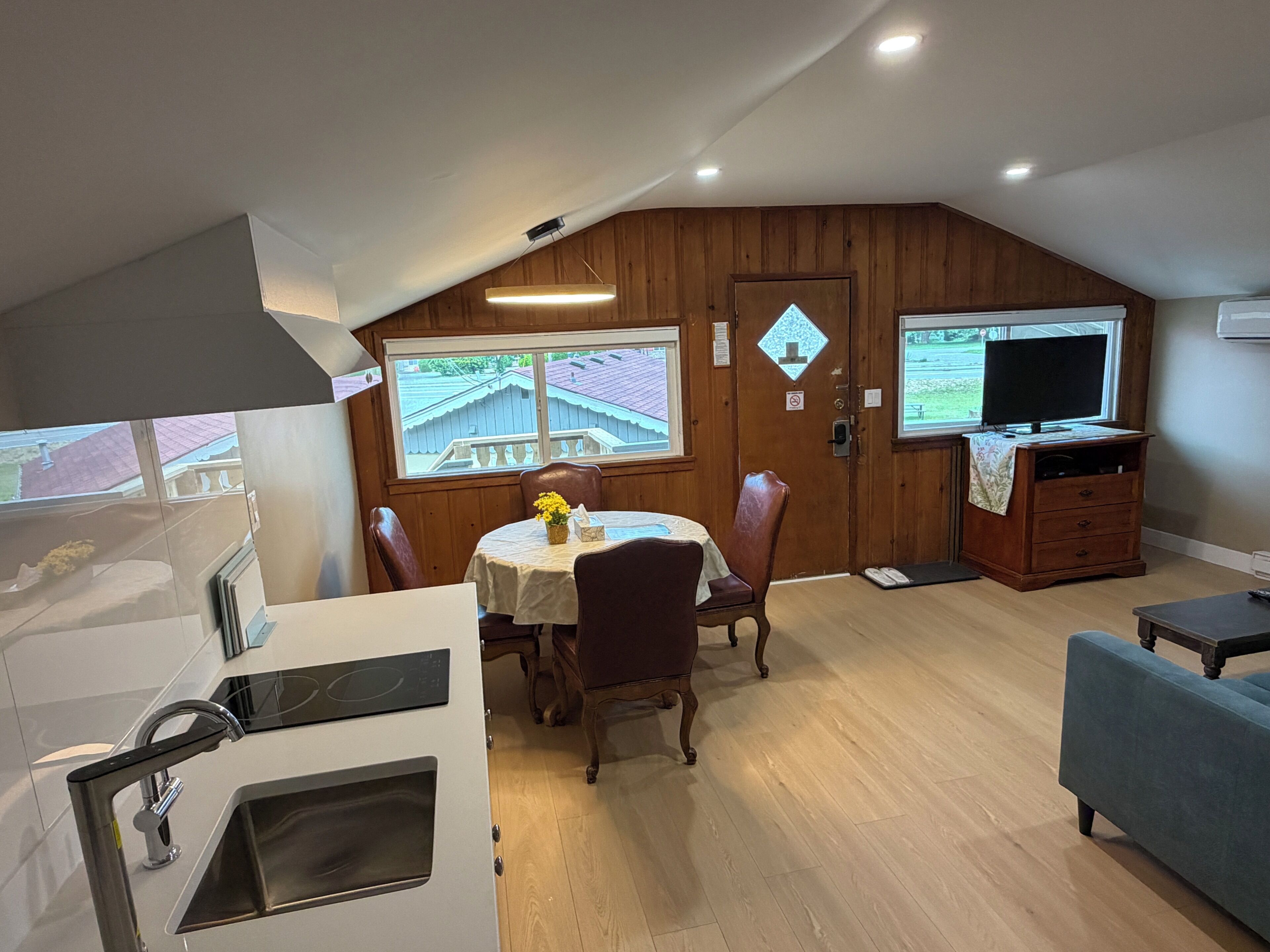 A compact kitchen with a built-in oven and sink, a dining area with a table and chairs, a television on a stand, and a view of a residential neighborhood through a window.