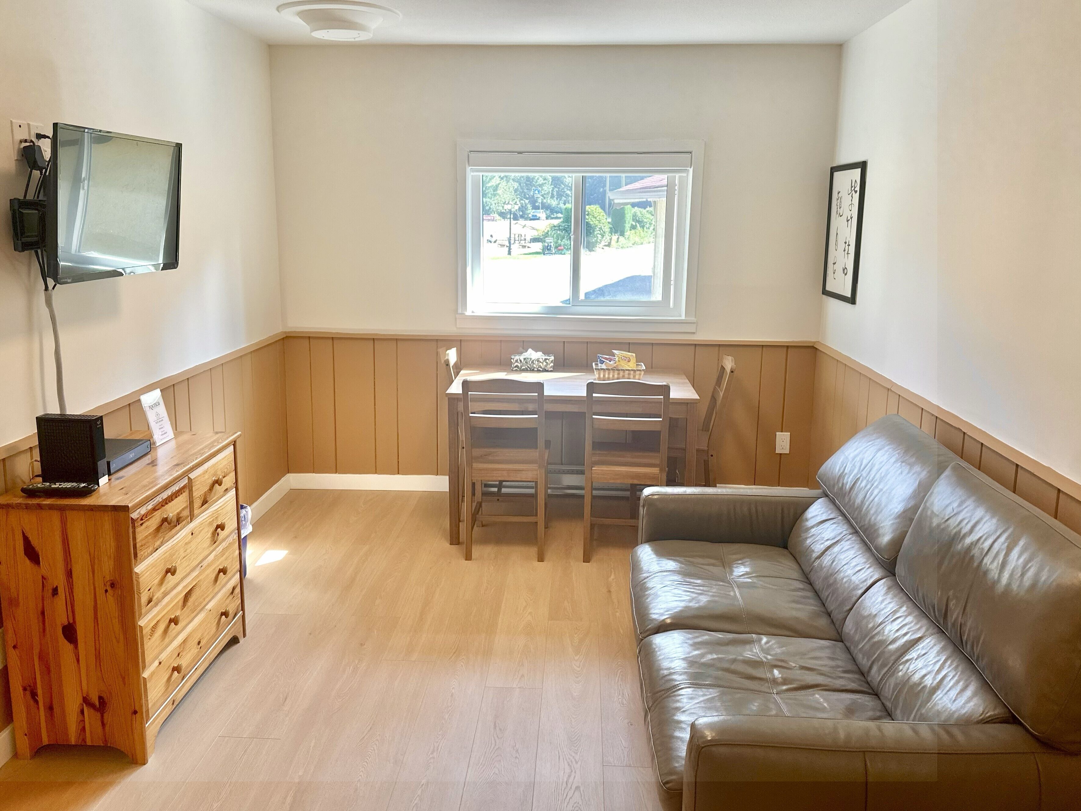 Un espacio habitable funcional con un aparador de madera, un sofá de cuero, una mesa de comedor con sillas, un televisor montado en la pared y una ventana con vistas al exterior.