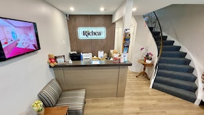 A reception area with a sign for Richca Chalets Resort, a sofa, a small table, a staircase, and a television displaying a room. - Richca Chalets Resort (Hope)
