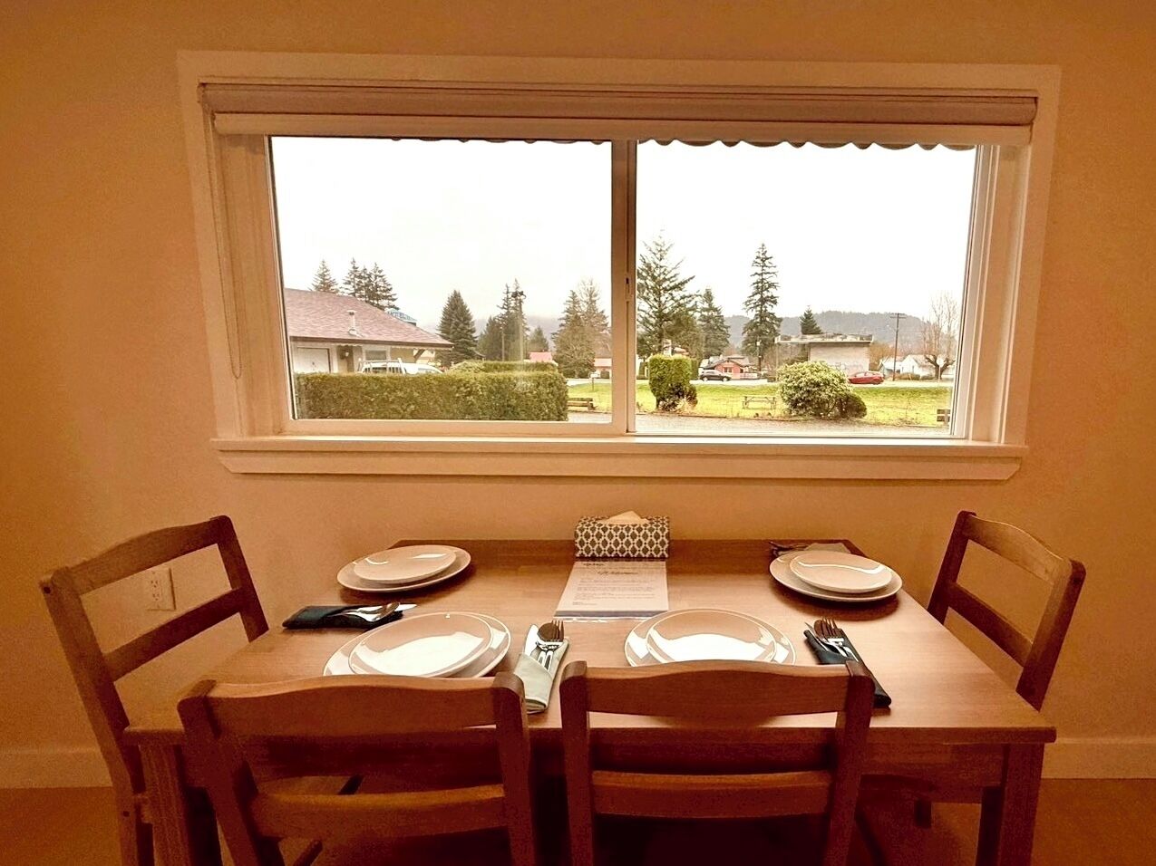 A dining table set for four with chairs, a window with a view of houses and trees, and a menu on the table.