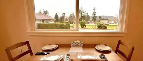 A dining table set for four with chairs, a window with a view of houses and trees, and a menu on the table.
