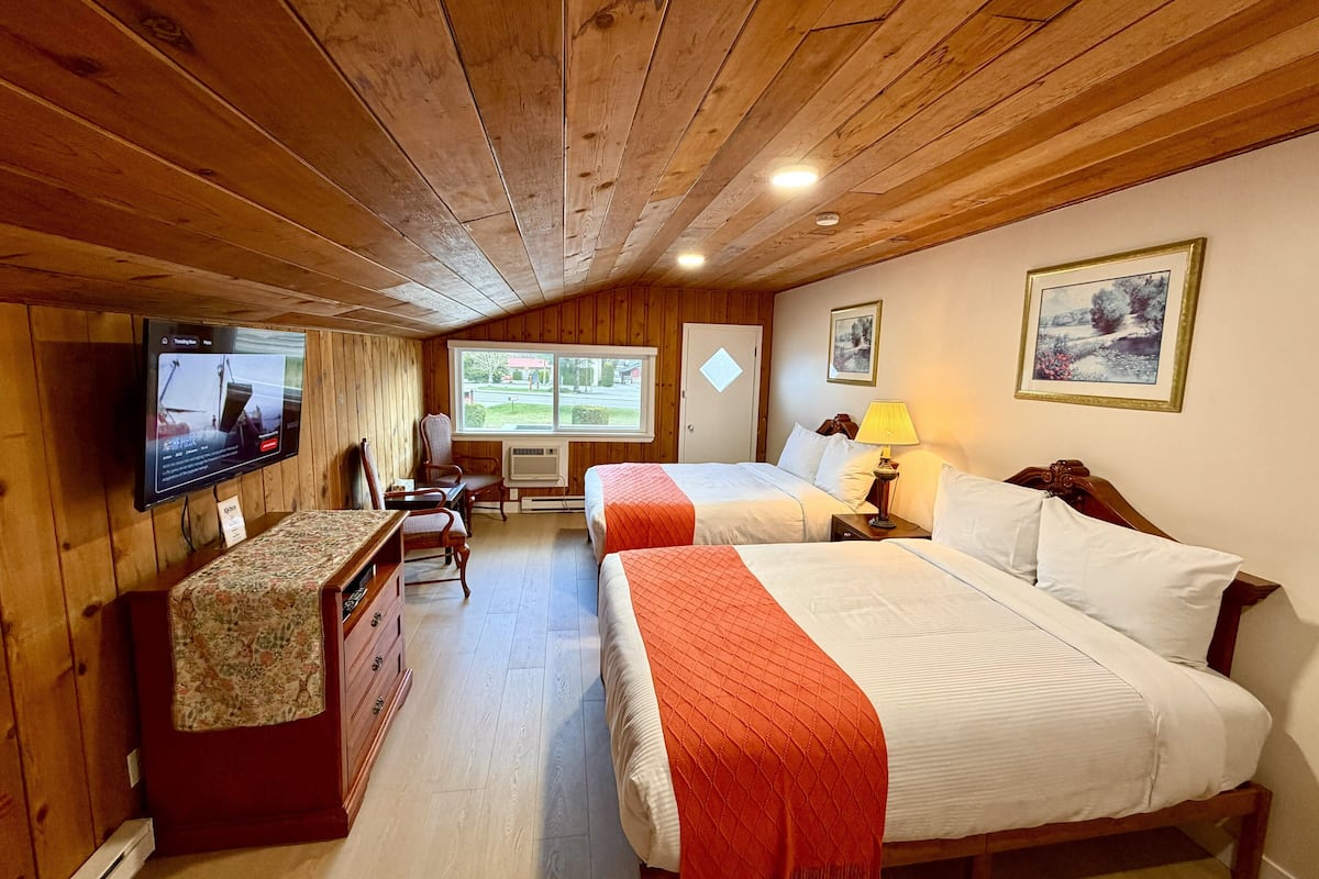 A room with wooden paneling, two beds with red and white bedding, a desk with a television, and a window with a view of greenery.