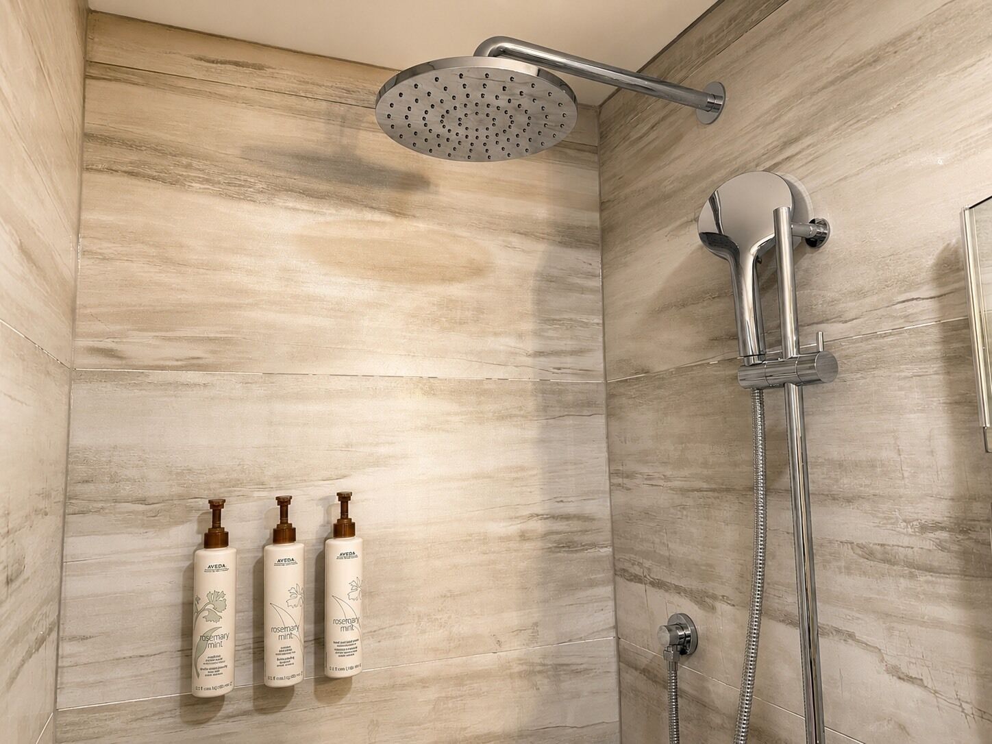 Une douche équipée d’un pommeau de douche à effet pluie et d’un pommeau de douche à main, le tout fixé sur un mur dont le revêtement imite le marbre.
