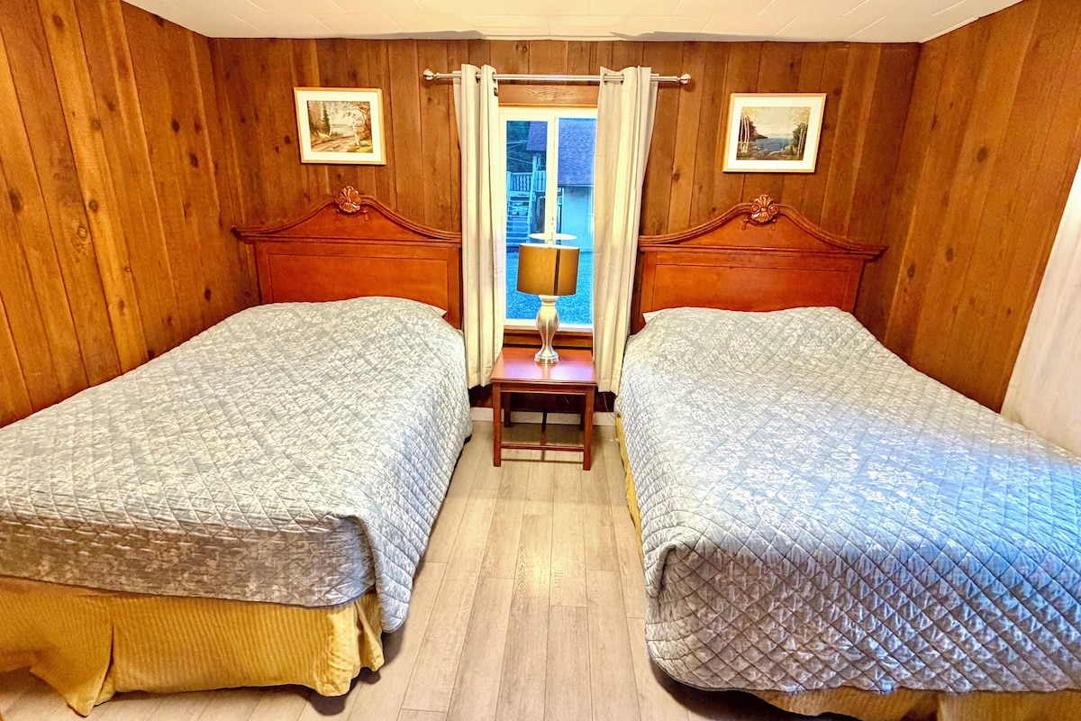 Una habitación con dos camas, paredes de madera, una ventana con cortinas y una mesita con lámpara.