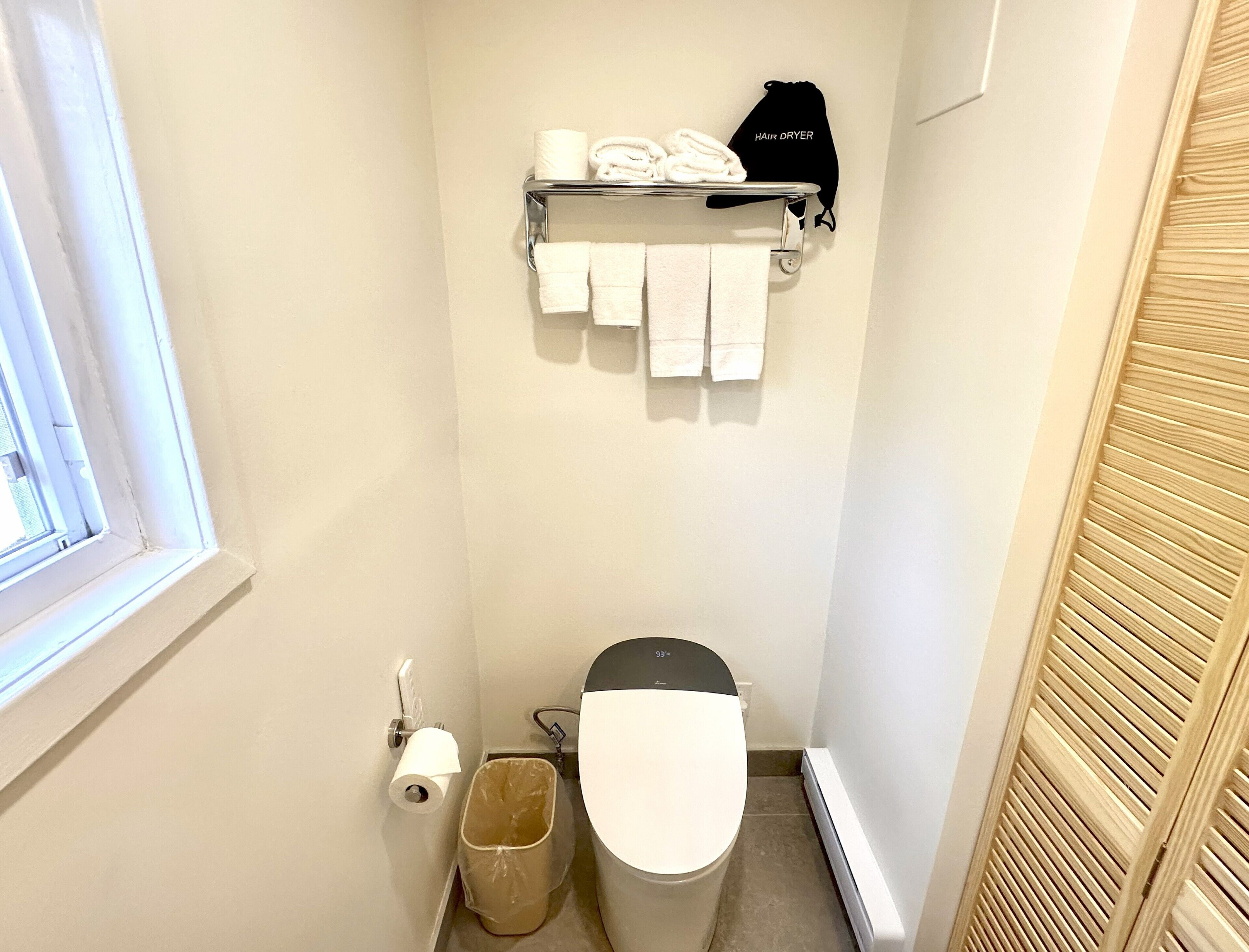 A small bathroom with a toilet, a shelf with towels and a hair dryer, a trash bin, and a window.
