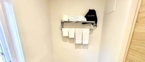 A small bathroom with a toilet, a shelf with towels and a hair dryer, a trash bin, and a window.