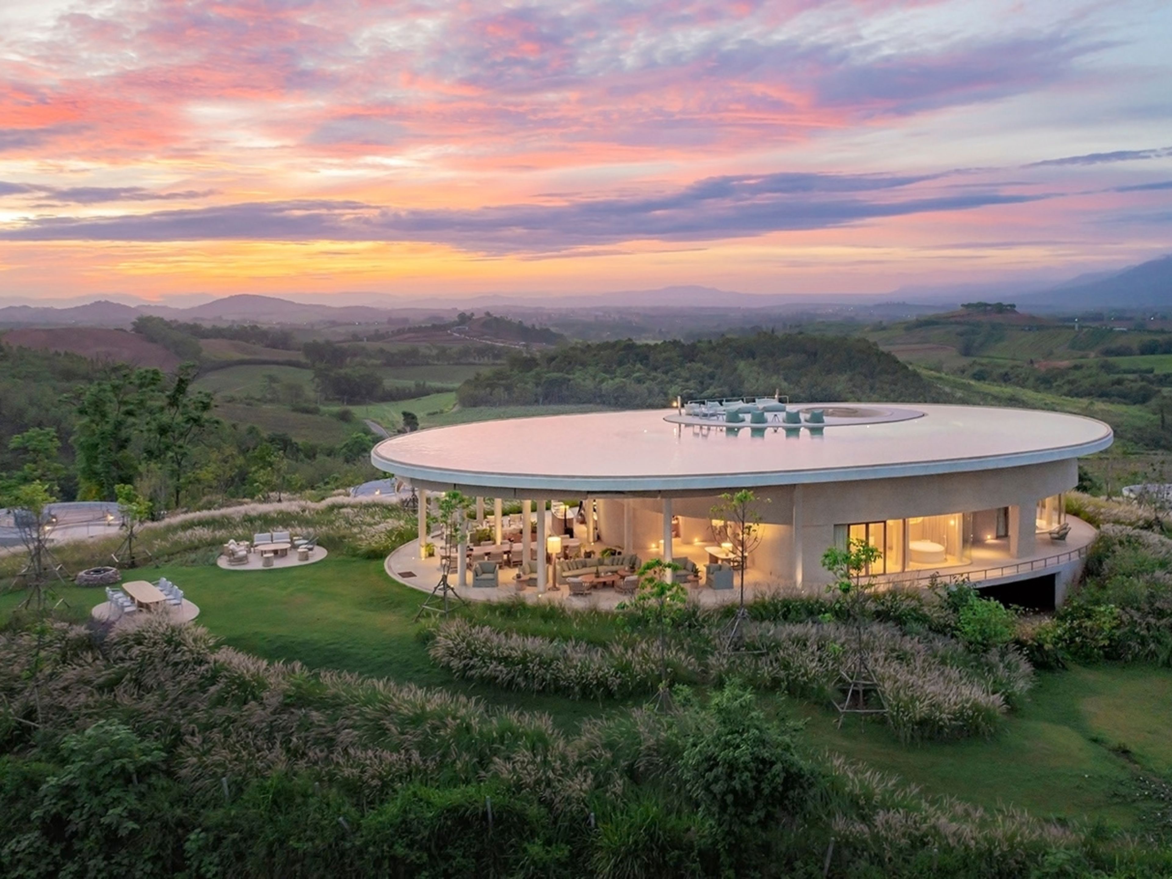 A modern circular building with a covered outdoor area, surrounded by lush greenery and hills in the background.