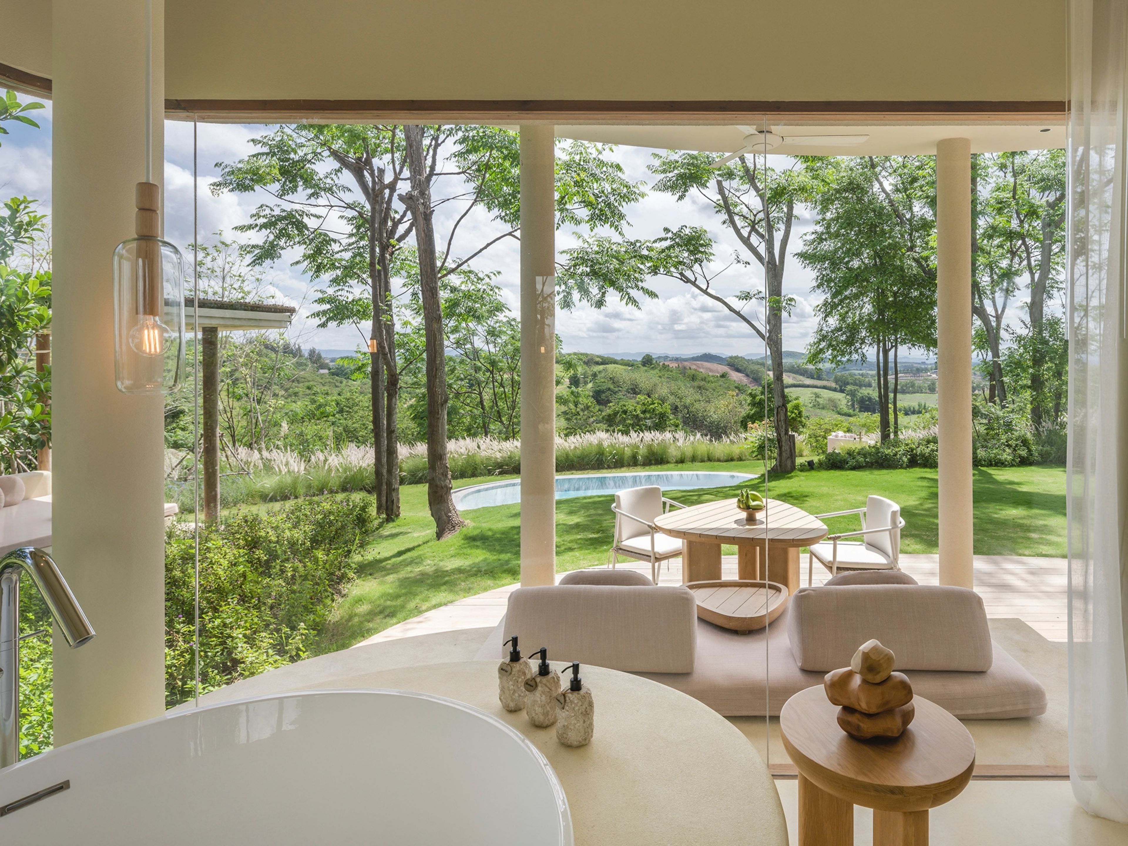 A modern bathroom with a large bathtub, a round table with chairs, and a view of a lush garden and pool.