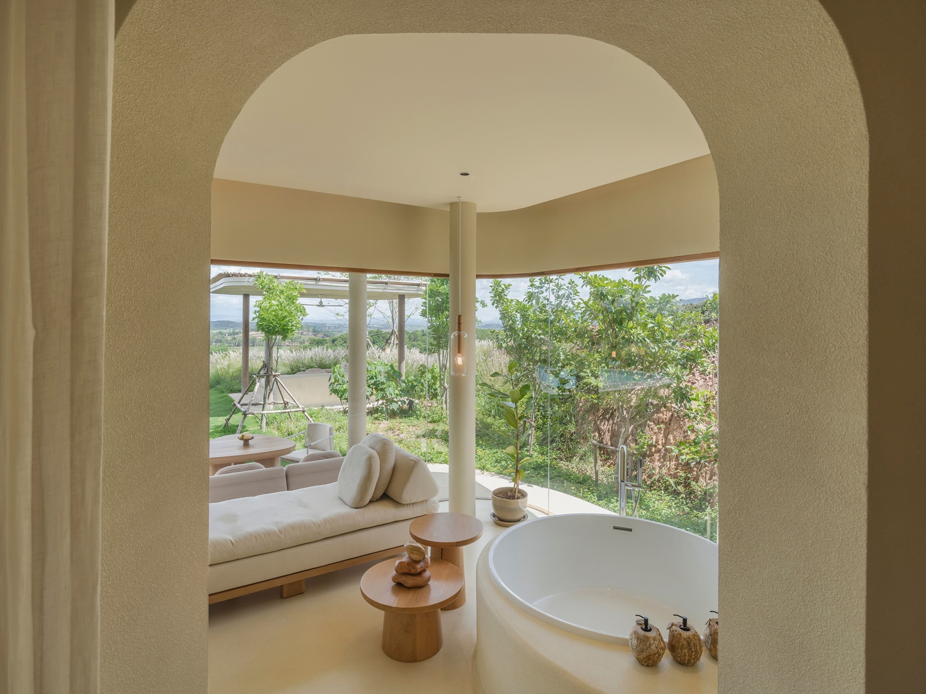 A modern bathroom with a freestanding bathtub, a sofa, and a view of a lush landscape.