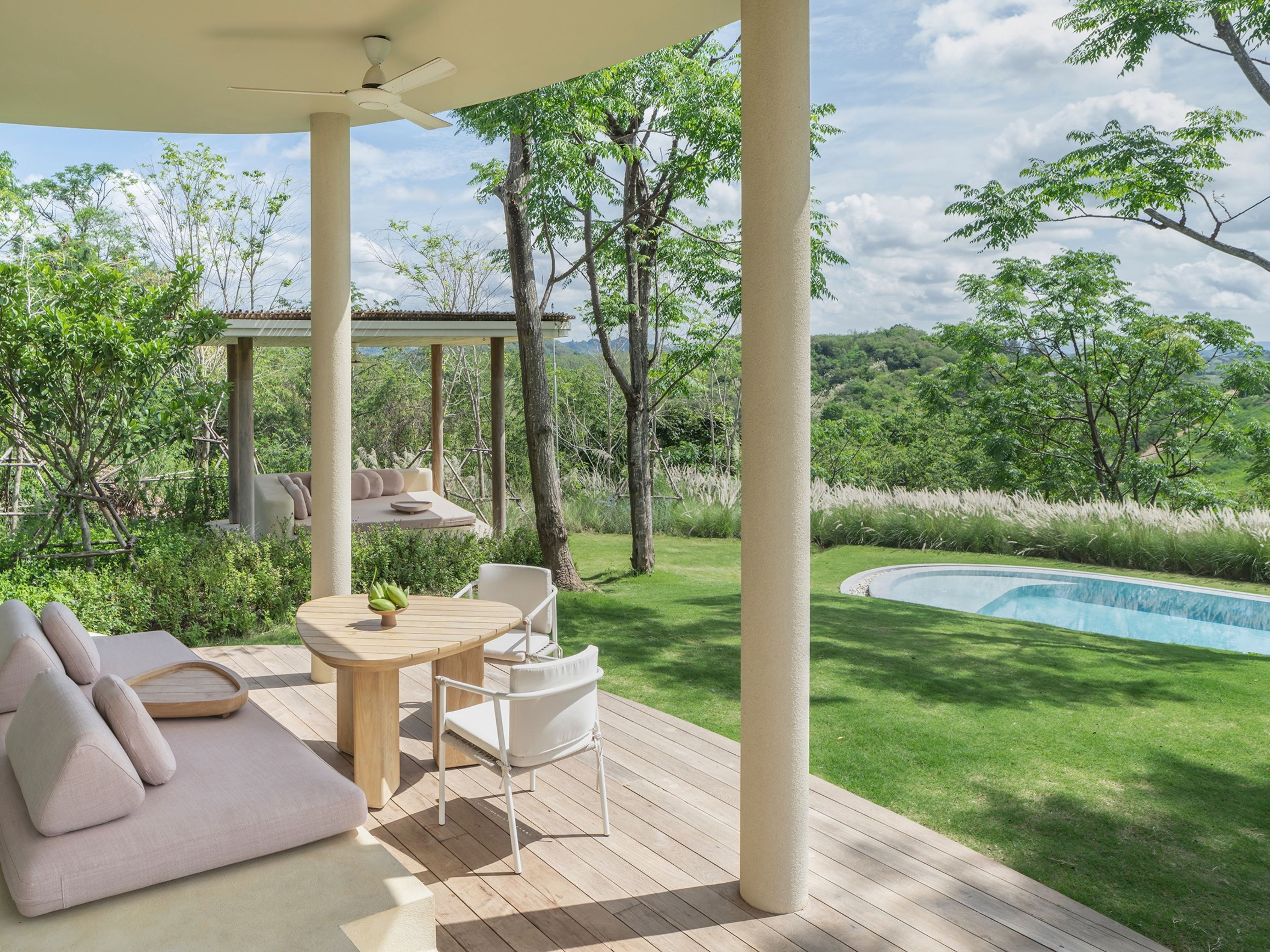 Un espacio de estar al aire libre techado, con piscina y abundante vegetación al fondo.