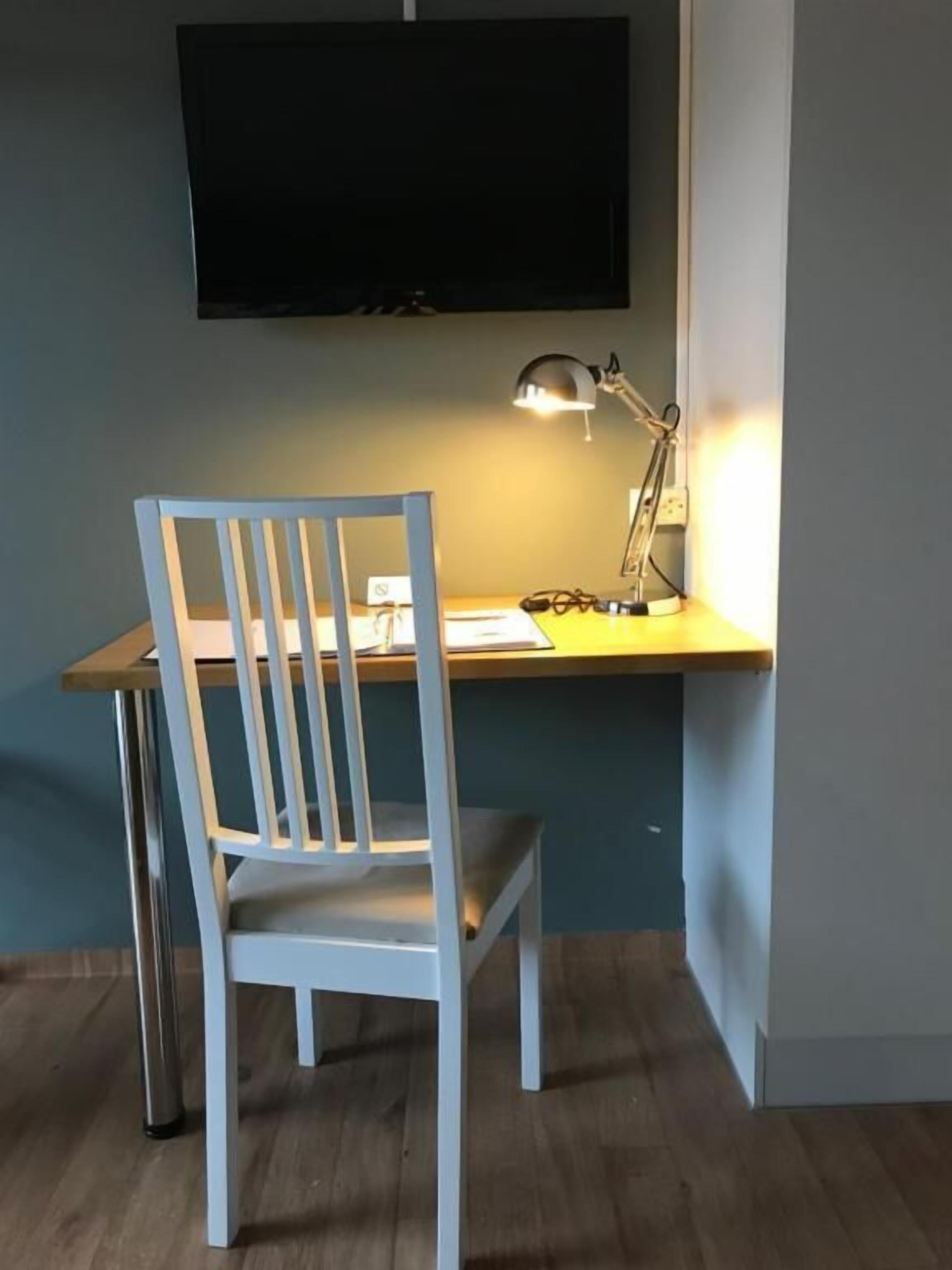 Una habitación con escritorio, silla, televisor montado en la pared y lámpara de escritorio.