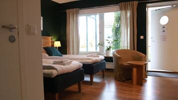 A hotel room with two beds, a chair, a small table, a lamp, and a window with curtains.