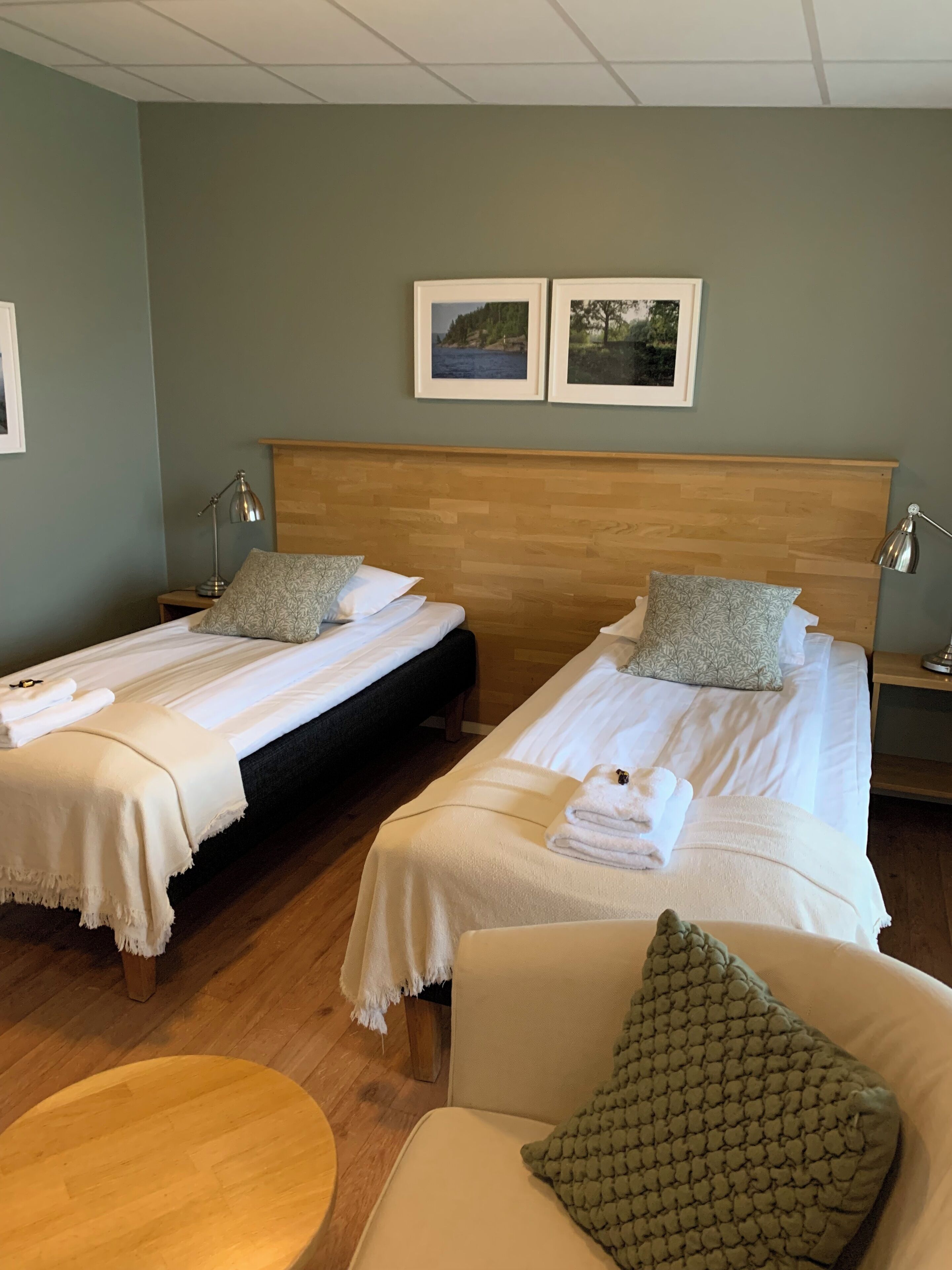 A hotel room with two beds, each with a wooden headboard, white linens, and a green pillow. There are two bedside tables with lamps, and a round wooden table in the center.