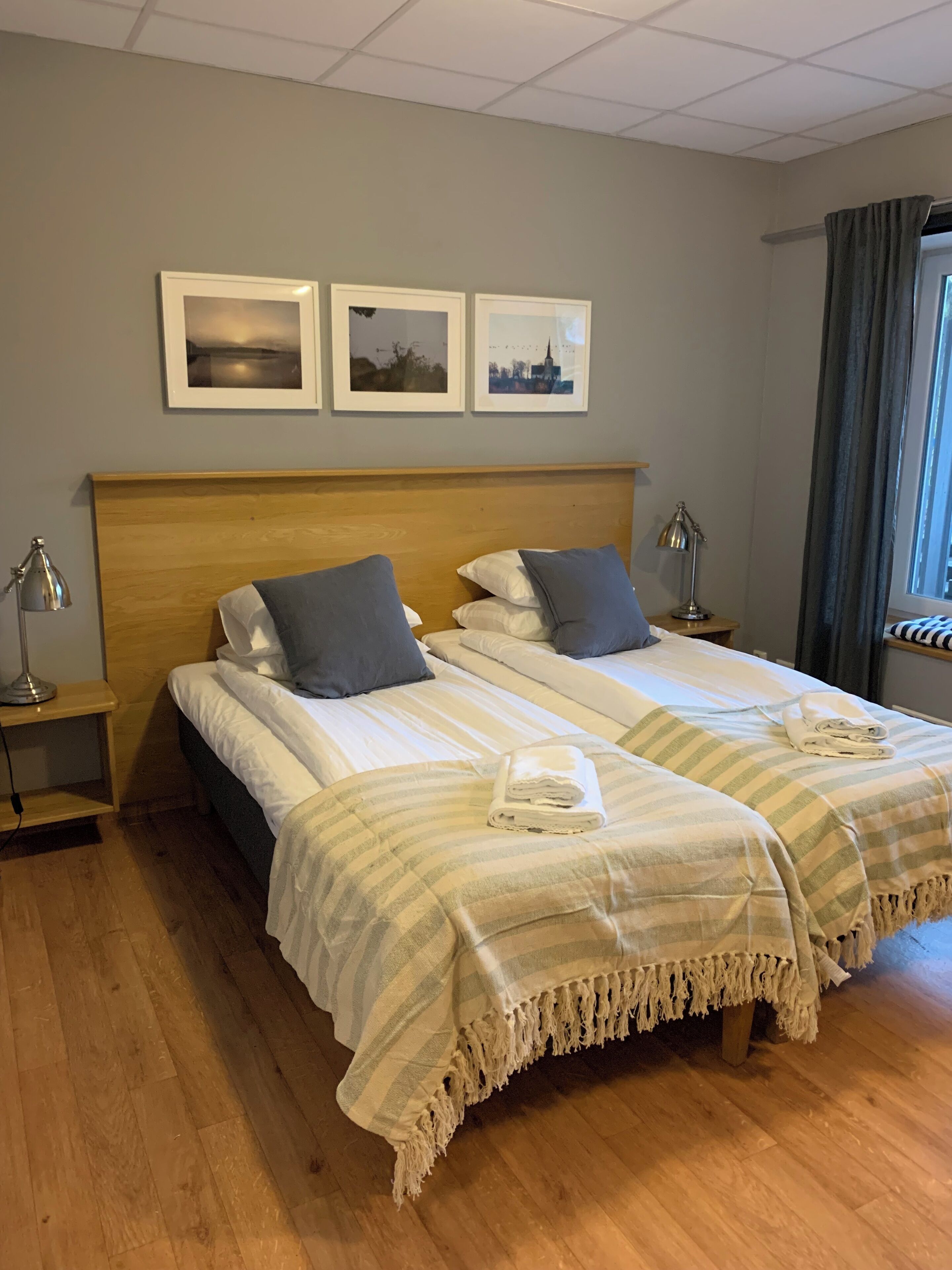 Una cama doble con cabecera de madera, dos mesitas de noche con lámparas y una ventana con cortinas.